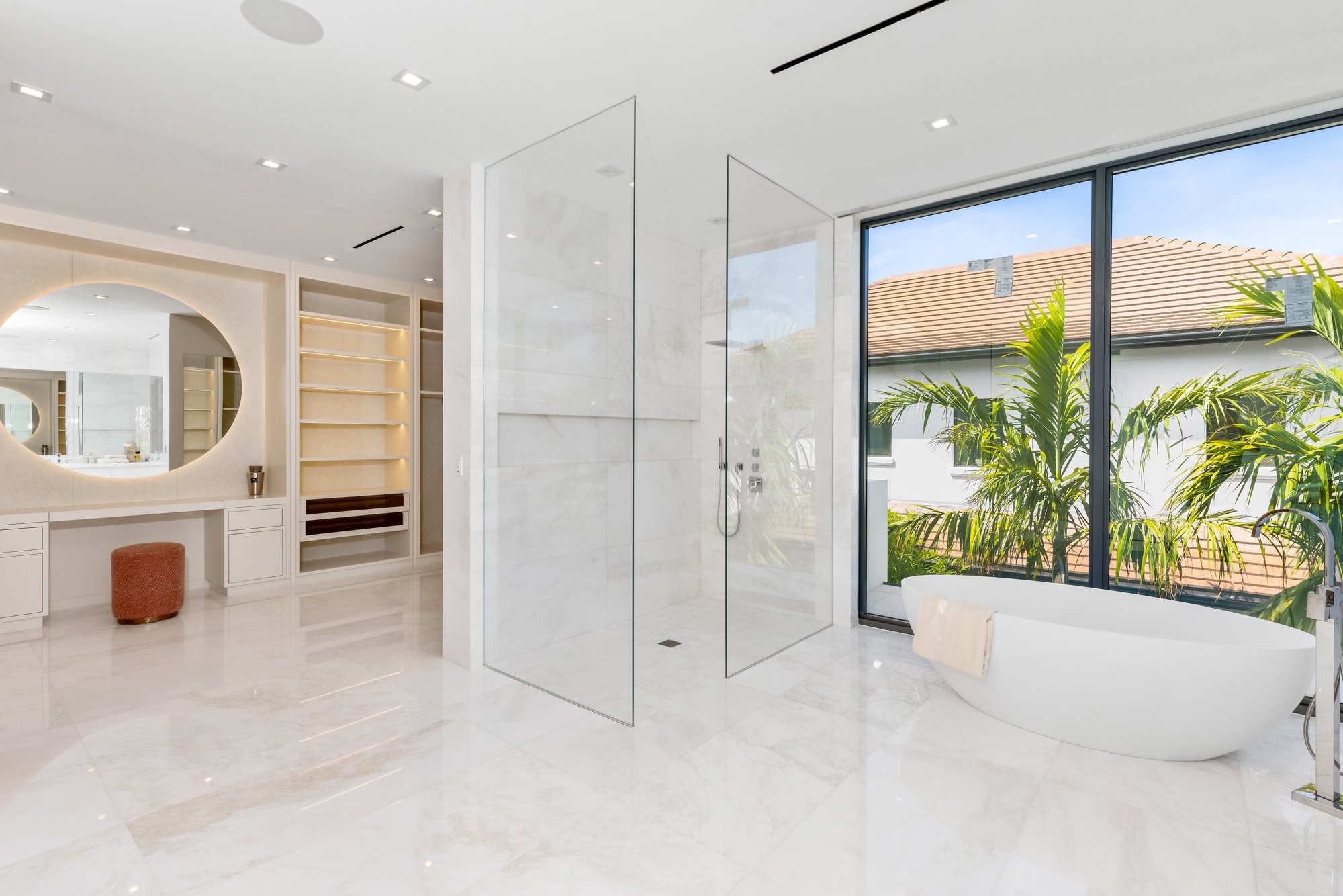 Modern bathroom with marble floors, a large walk-in shower, a freestanding bathtub, and a large window with a view of outdoor plants and houses.