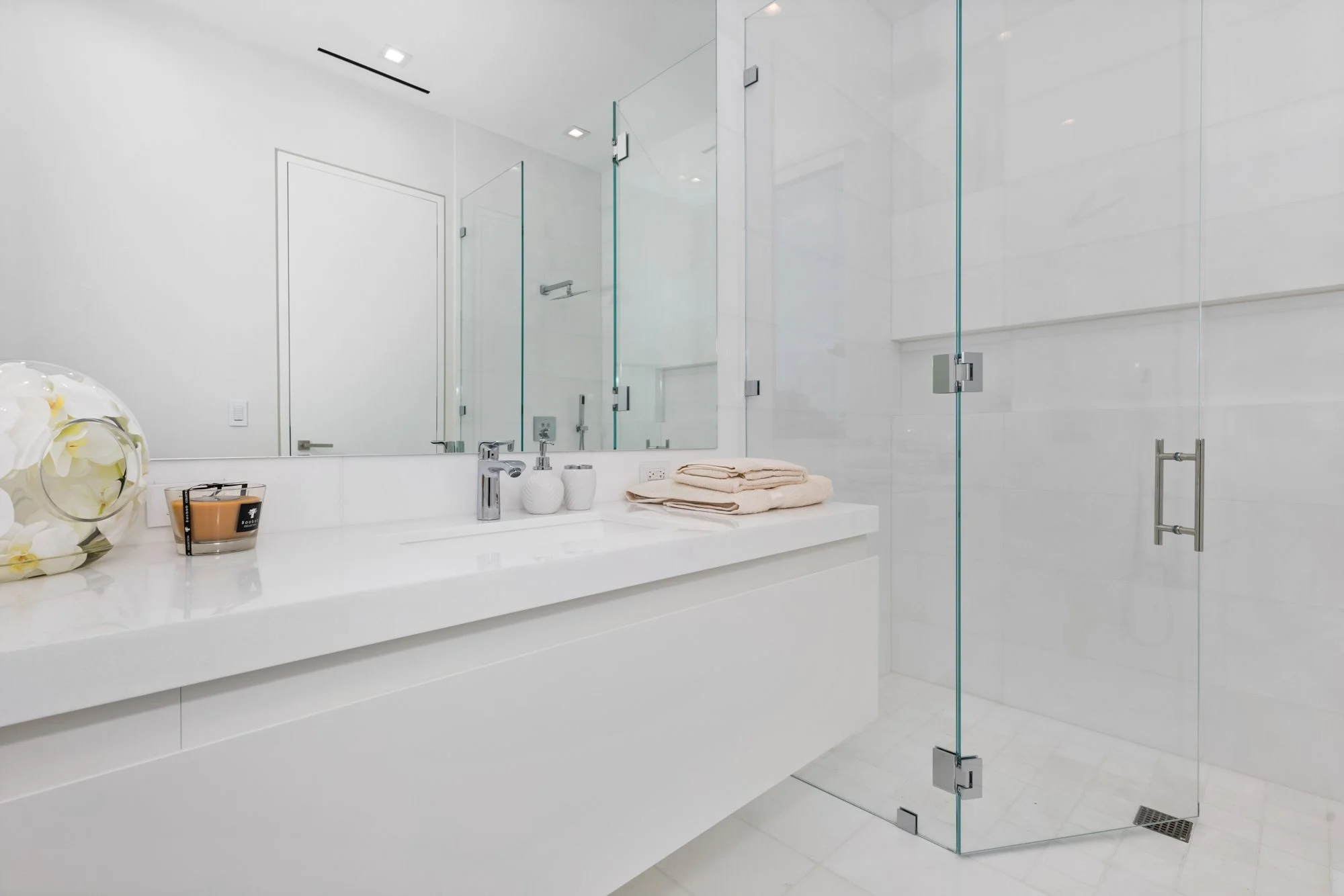 Modern white bathroom with a large mirror, white countertop with folded towels, a small flower arrangement, and a glass-enclosed shower.