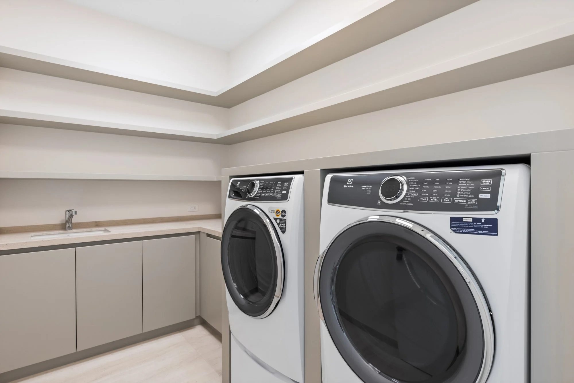 Laundry room with beige cabinets, beige countertop, front-loading washing machine and dryer, small sink with faucet, and beige walls with a pamphlet on the washing machine.