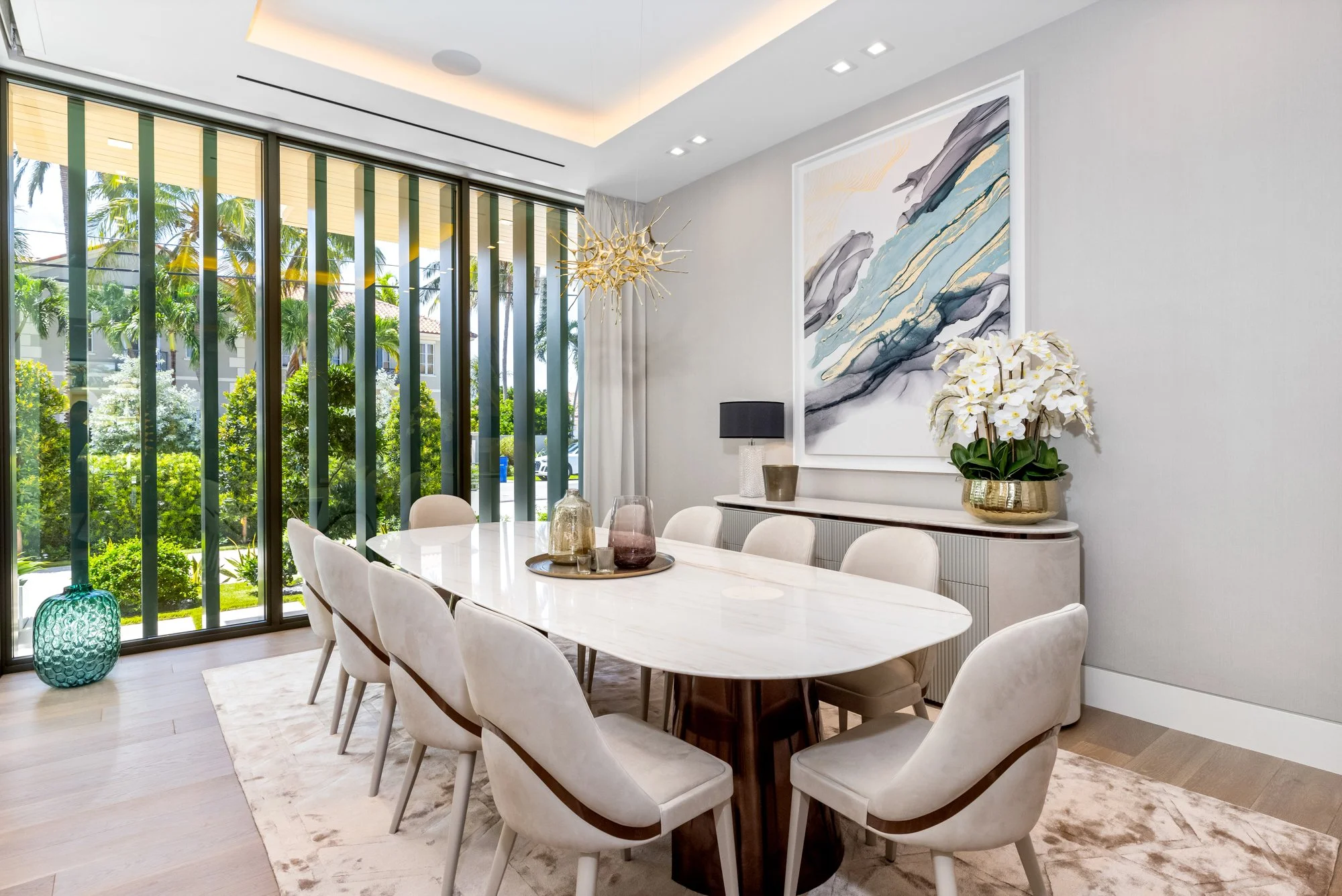 Modern dining room with a white marble table, nine beige upholstered chairs, and a large abstract painting on the wall. Large glass windows show greenery outside. Decor includes a gold chandelier, vases, and a potted orchid.