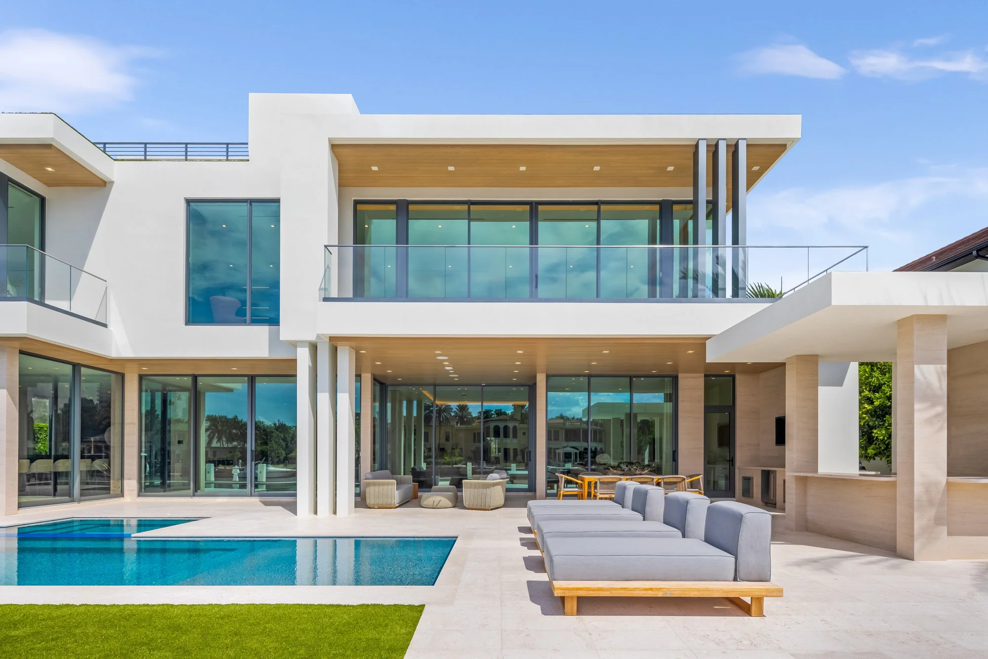 Modern multi-story house with large glass windows, a swimming pool, outdoor seating area, and balcony. Bright and sunny day with blue sky.