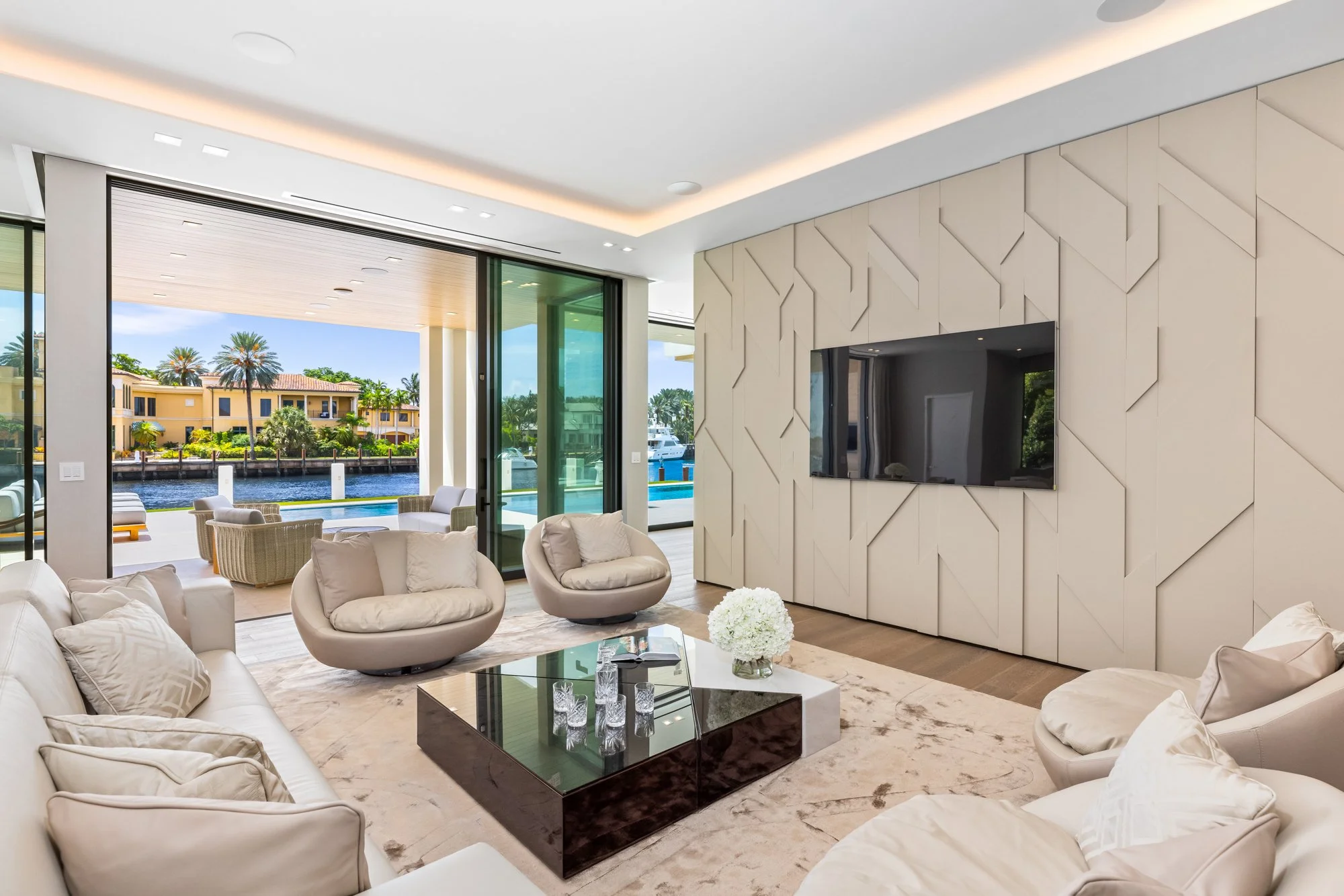 Modern living room with beige sofas and chairs, glass coffee table with flowers and glasses, large flat-screen TV on decorated wall, sliding glass doors leading to outdoor patio with water view and pool.