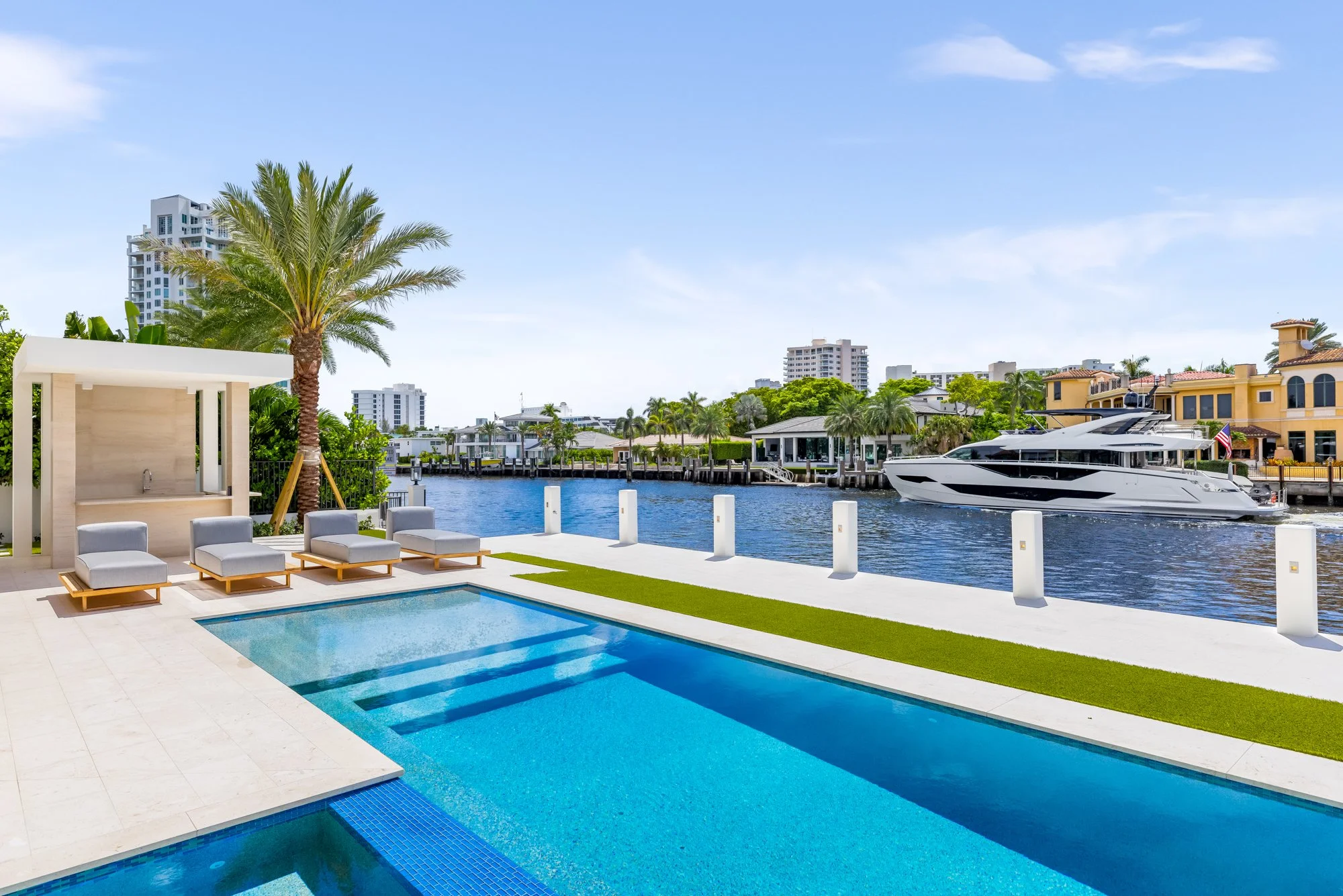 Luxury waterfront house featuring a swimming pool, sun loungers, palm trees, a dock with a yacht, and city buildings in the background.