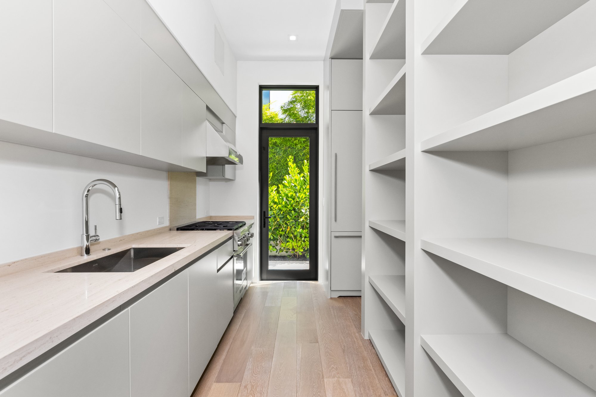 Modern white kitchen with beige countertop, black door with glass panel, open shelves, wooden floor, and greenery outside the door.