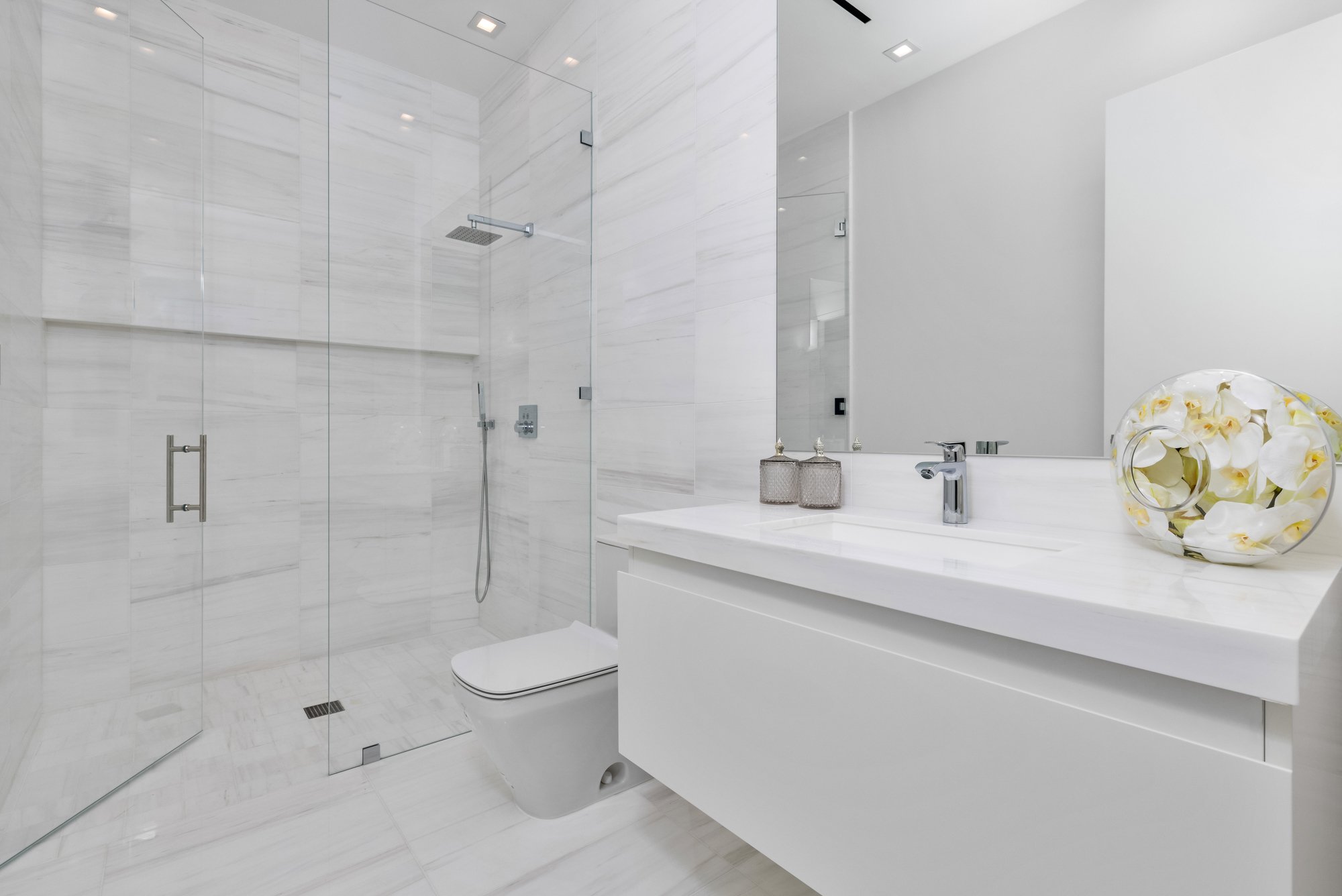 Modern bathroom with a glass-enclosed shower, white vanity with silver fixtures, a mirror, and a decorative vase with white flowers.