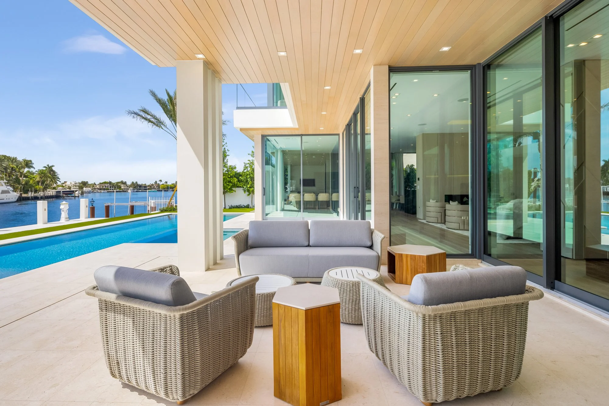 Modern outdoor patio with seating area, overlooking a swimming pool and waterfront with boats, featuring glass doors and windows, and a wood-paneled ceiling.