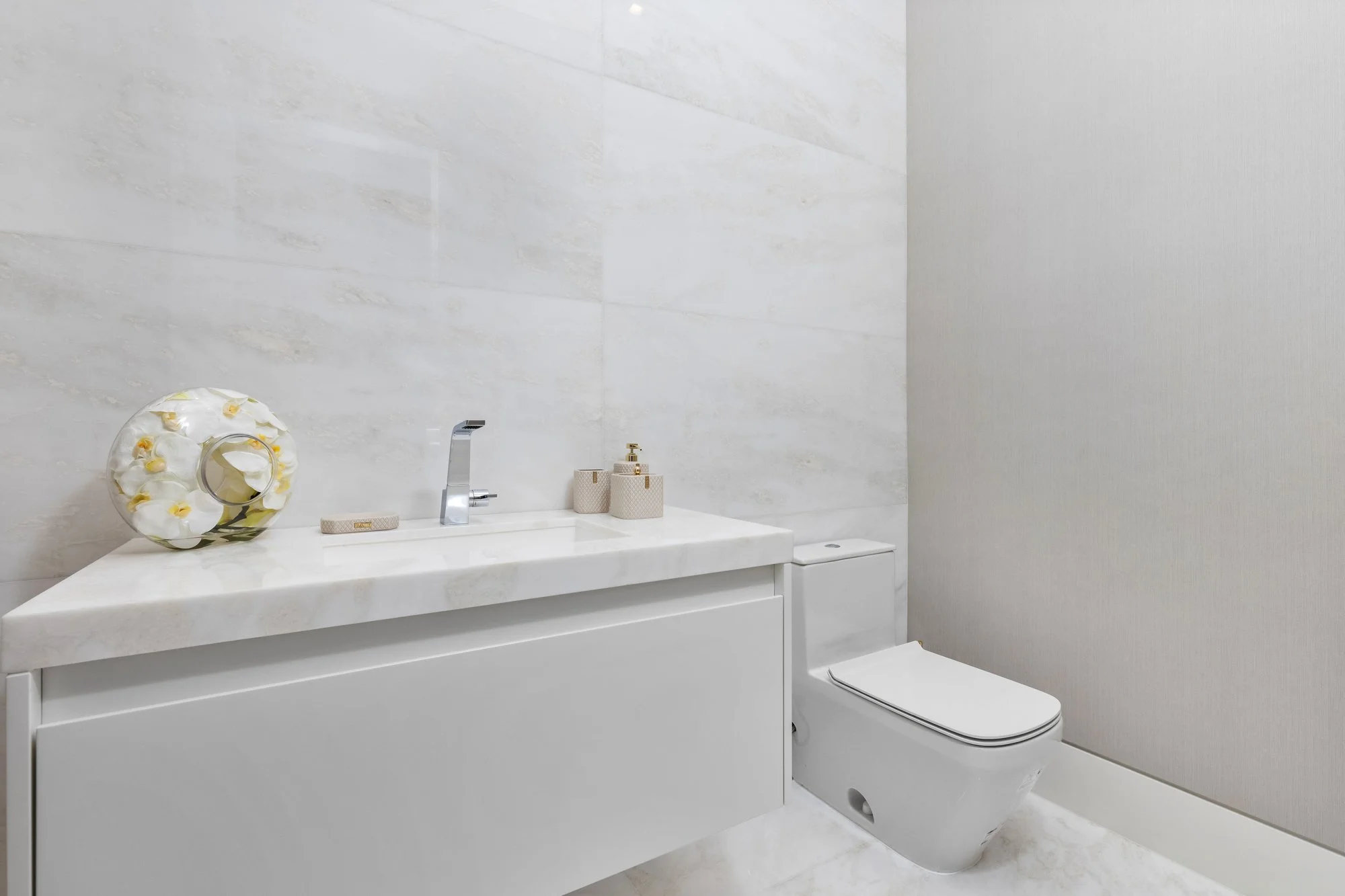 Modern bathroom with white marble sink vanity, decorative flower-shaped glass vase, soap dispenser, and toothbrush holder, and a wall-mounted toilet, in a minimalist design.