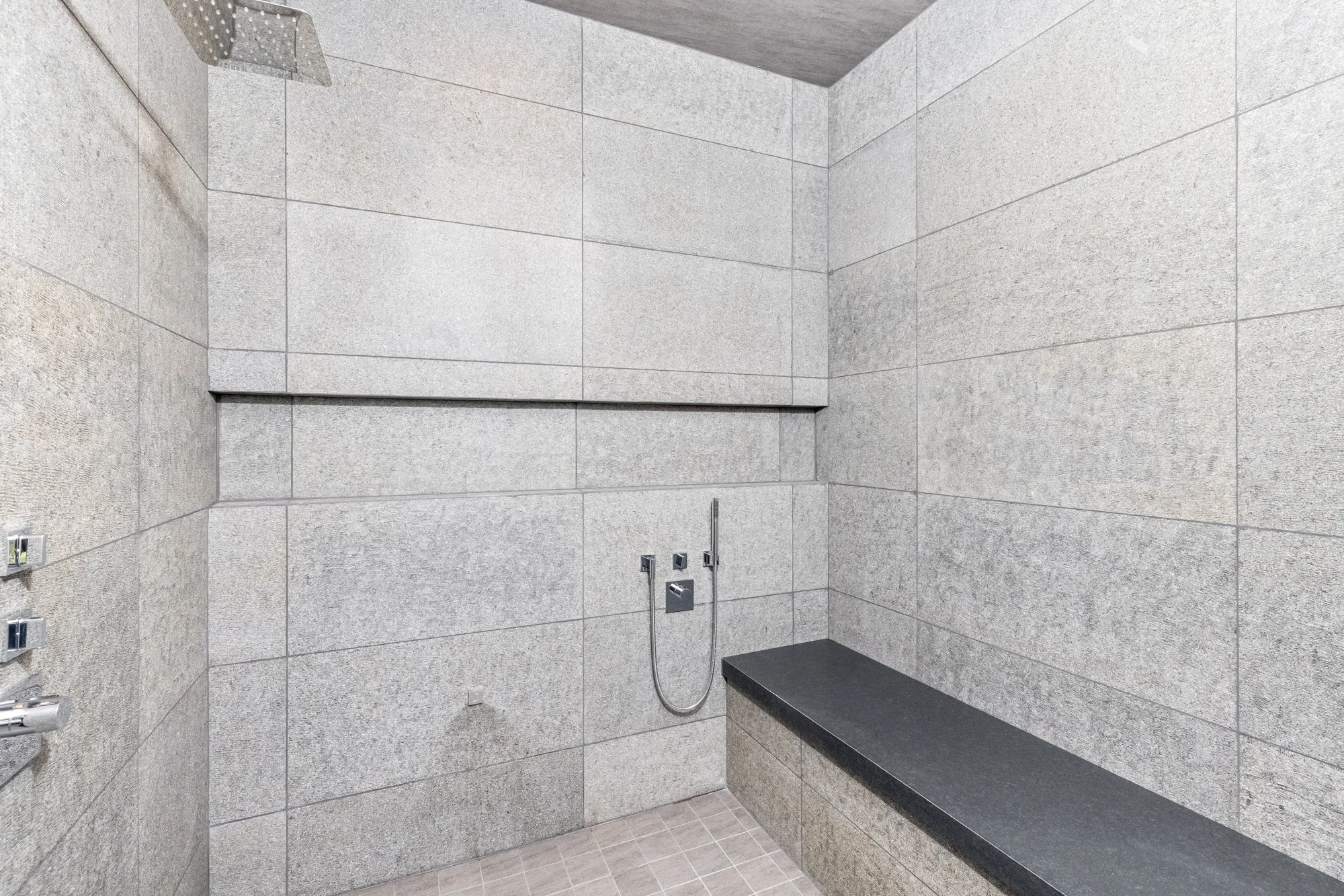 Modern shower area with gray tiled walls, a black bench, and a shower head.