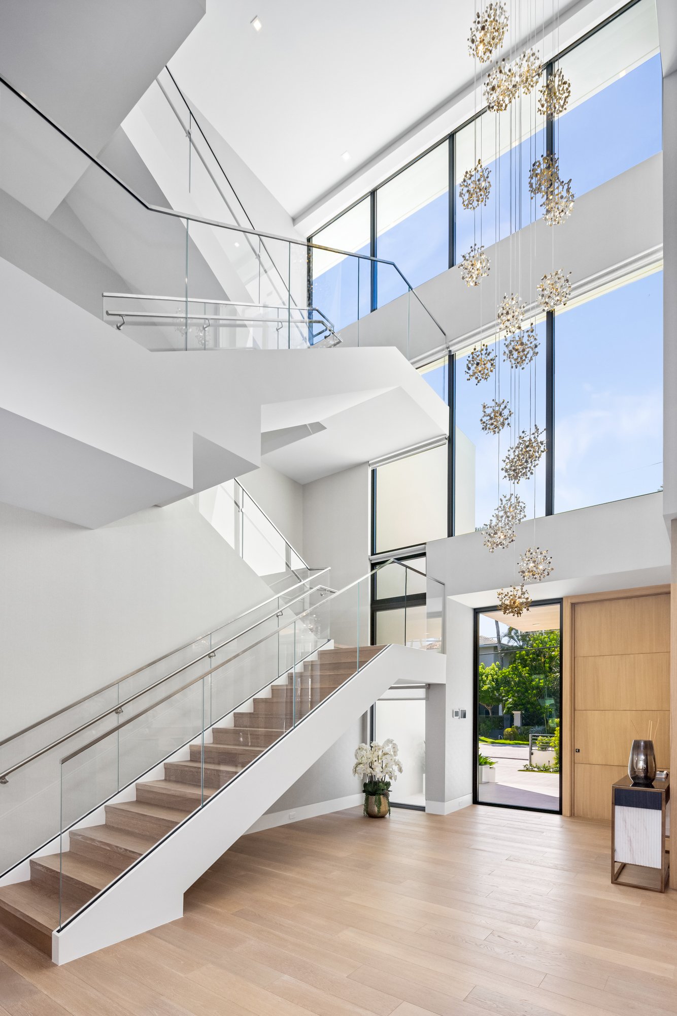 Interior view of a modern, minimalist house with a staircase, large windows, and a high ceiling feature with hanging decorative elements. There's a small table with a vase at the entrance, and a potted plant by the door, with sunlight illuminating th
