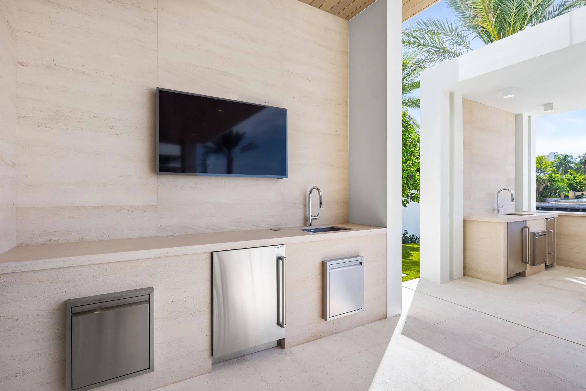 Outdoor kitchen area with built-in stainless steel appliances, a wall-mounted TV, sinks, and a view of palm trees and greenery.