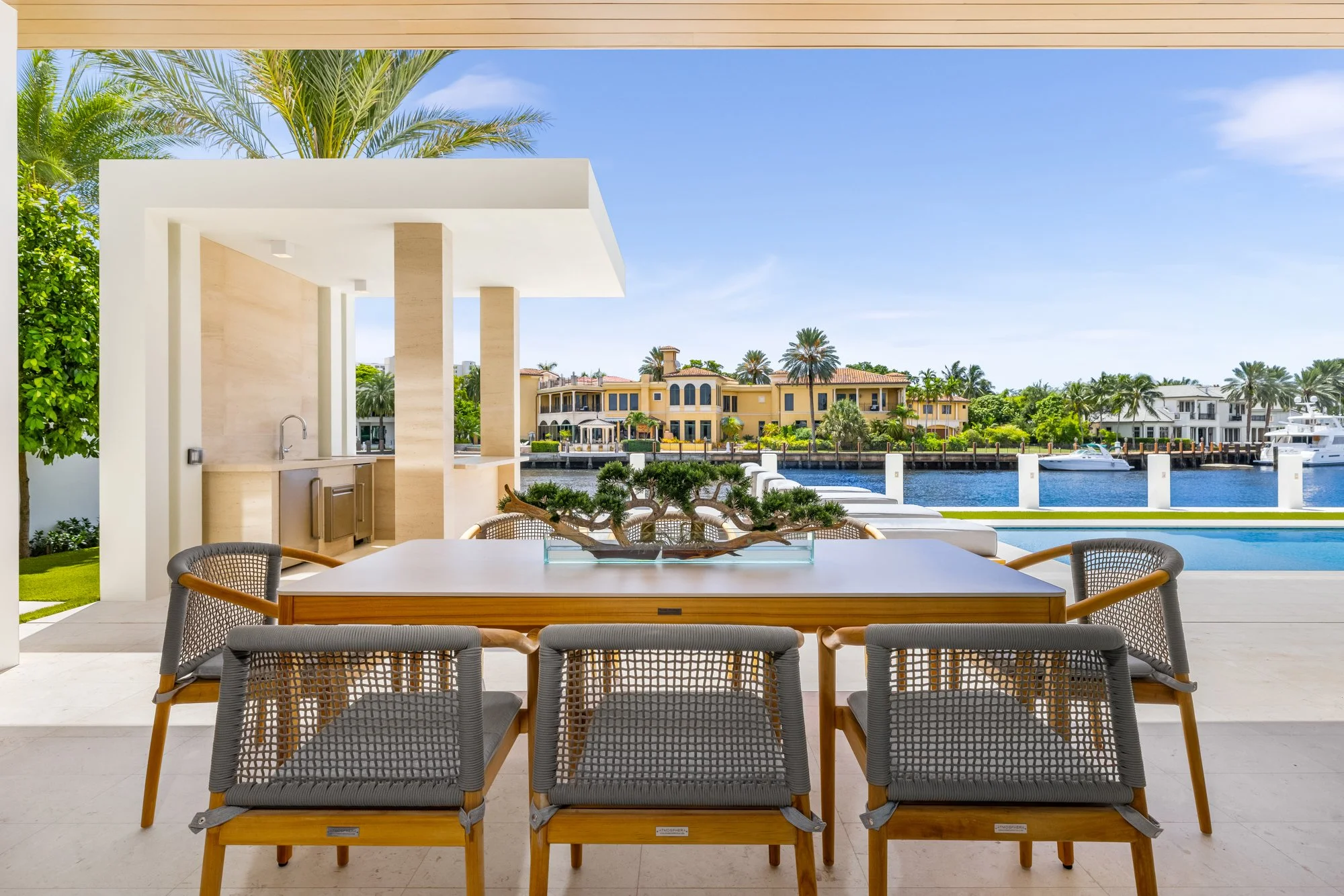 Outdoor dining area overlooking a waterway with yachts and luxurious houses, featuring a modern table with a bonsai tree centerpiece, surrounded by six chairs, under a partial roof, with lush greenery and palm trees in the background.