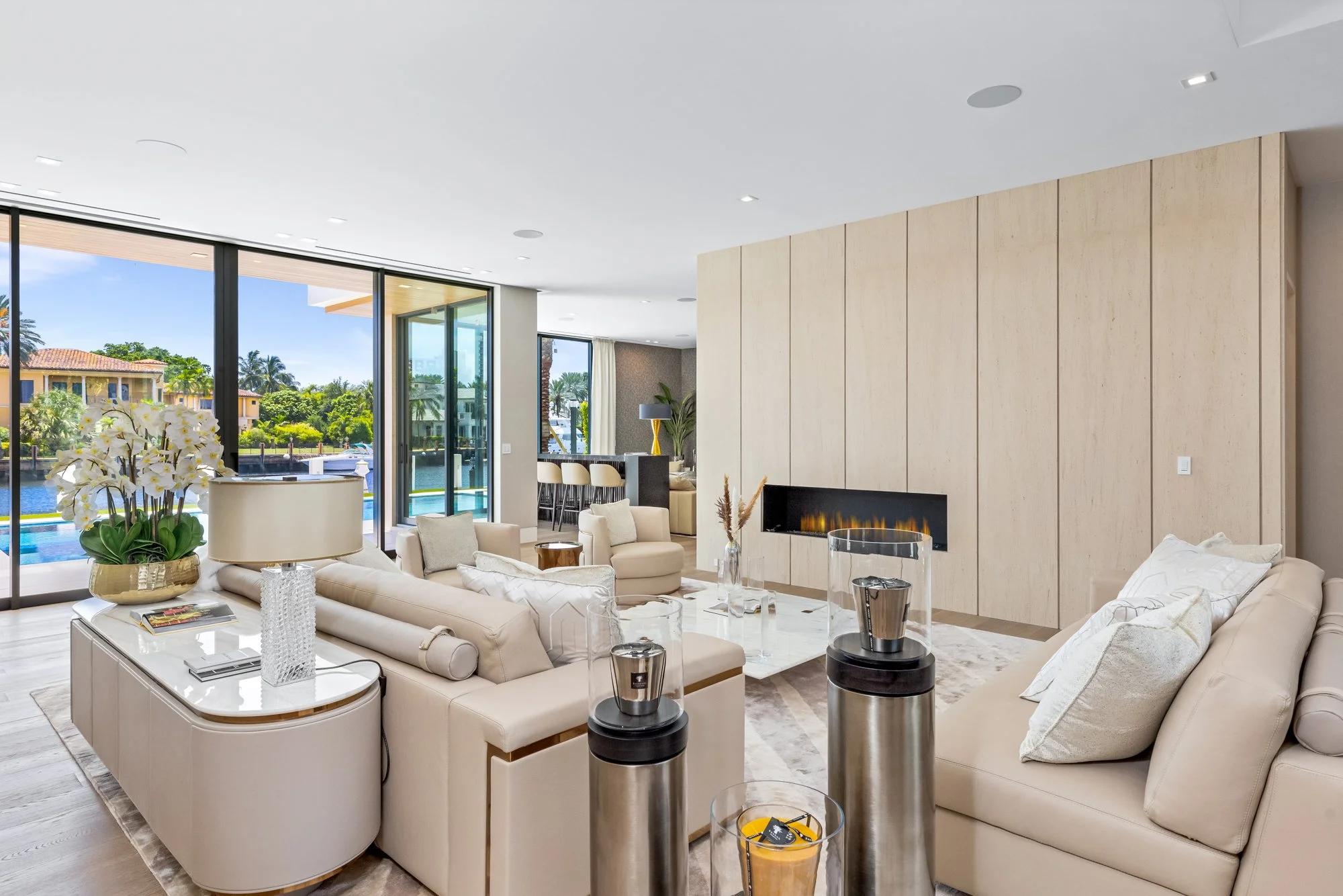 Luxurious modern living room with white leather sofas, a marble coffee table, an electric fireplace built into a wooden wall, large glass sliding doors leading to a patio with a pool, and tropical foliage outside.