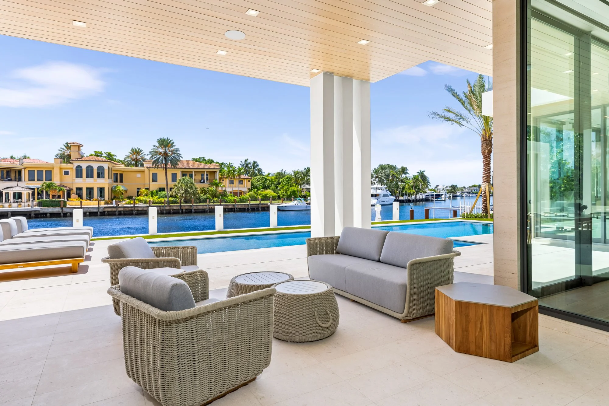 Modern outdoor patio with grey wicker chairs, a grey sofa, and a small wooden side table overlooking a swimming pool and a canal with boats and yellow houses under a clear blue sky.