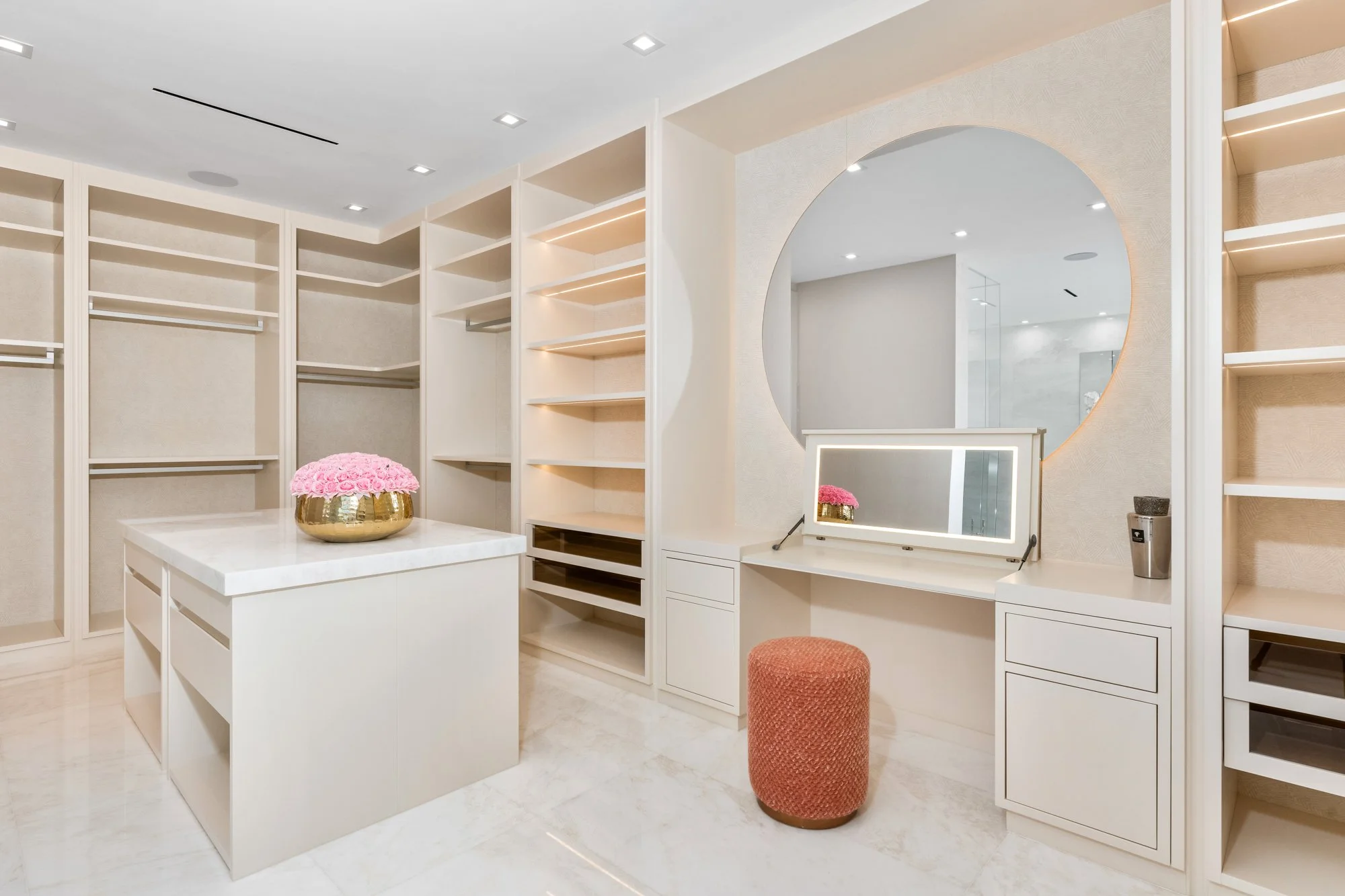 Modern walk-in closet with white shelving, a marble-topped island, pink flowers in a gold bowl, and a vanity with a round mirror.