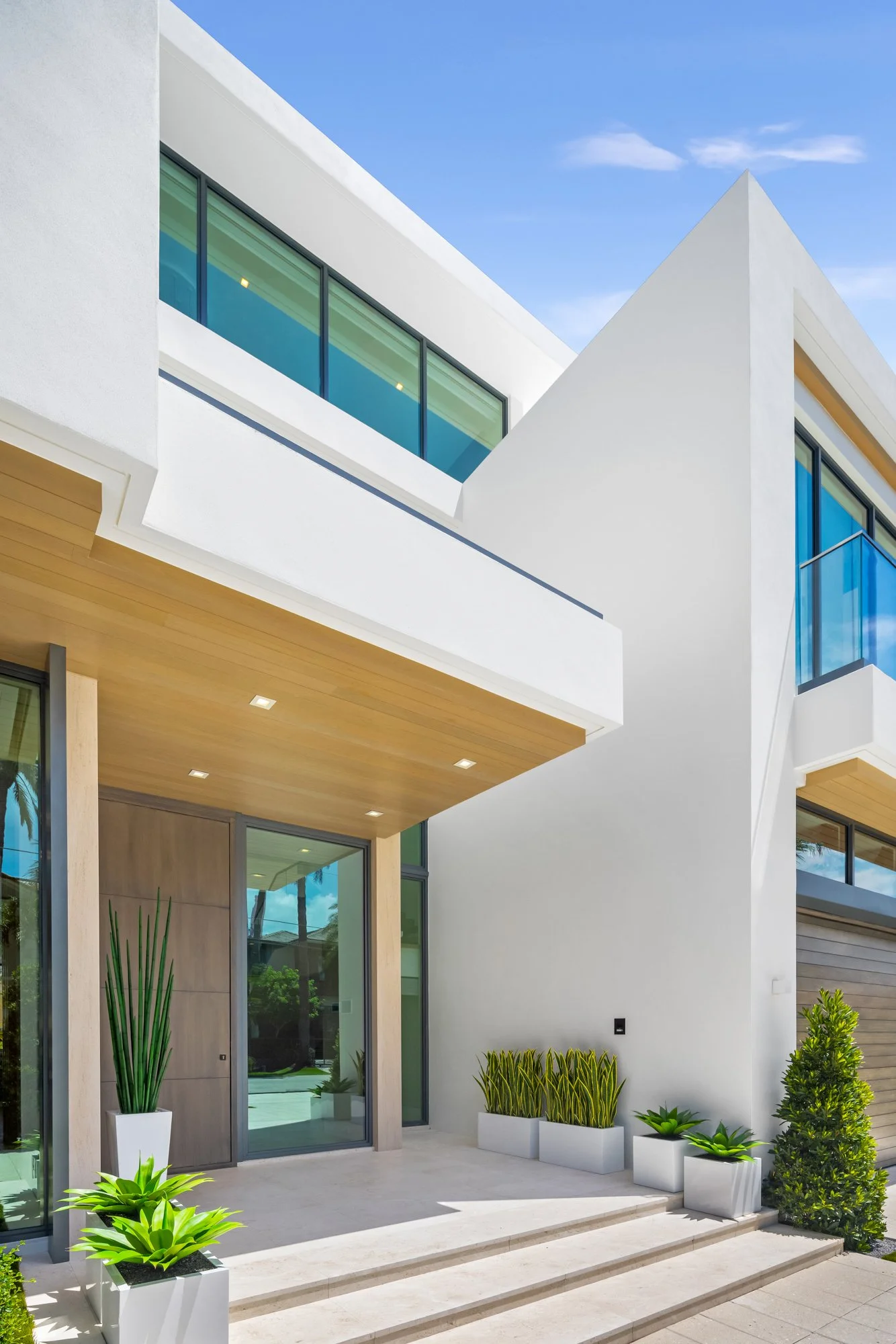Modern multi-story house with white exterior, wooden accents, and large glass windows, featuring potted plants at the front entrance.
