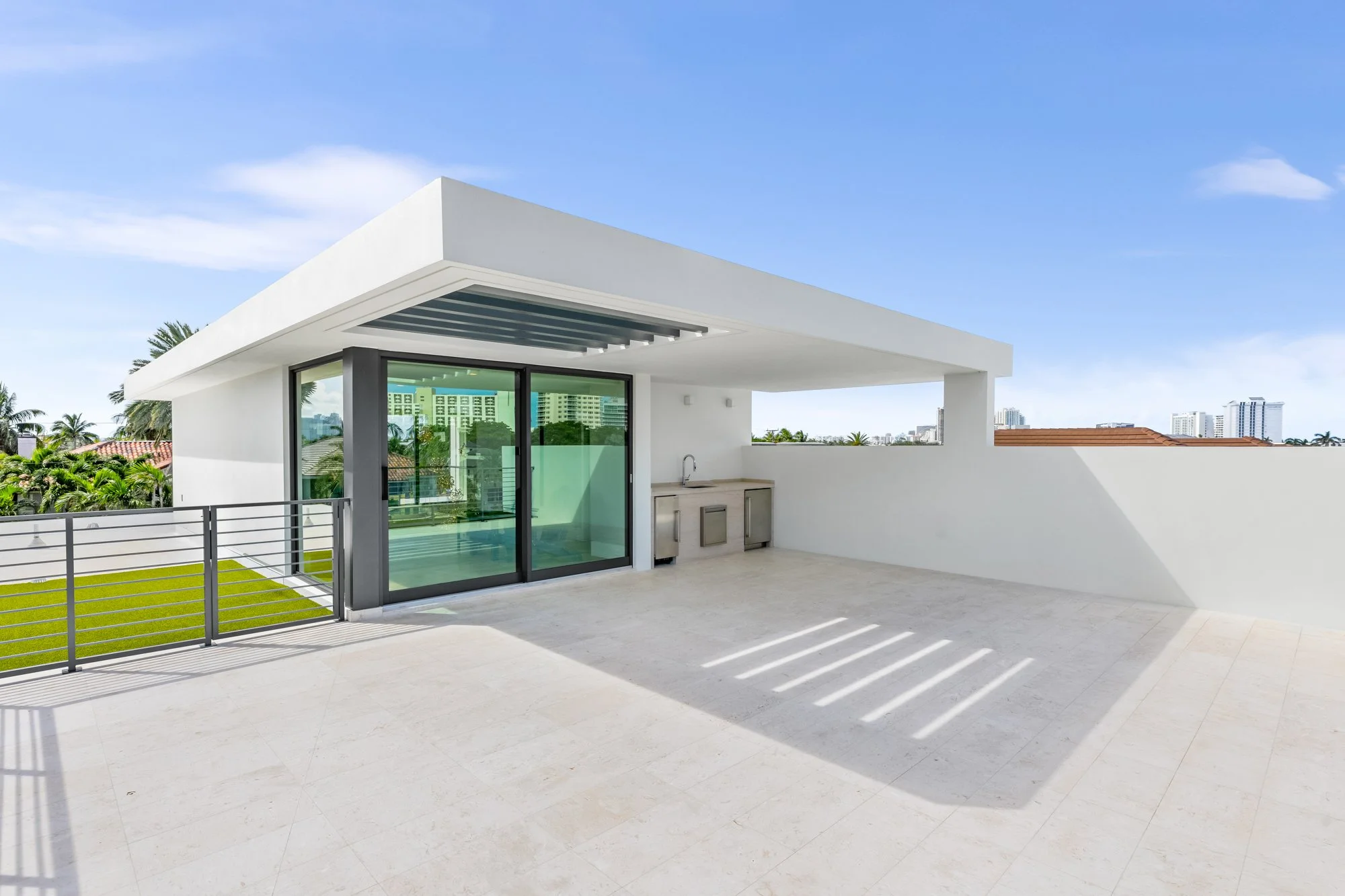 Modern rooftop terrace with white walls, glass sliding door, small kitchen area, and a partial view of city buildings and palm trees in the background, under a blue sky.