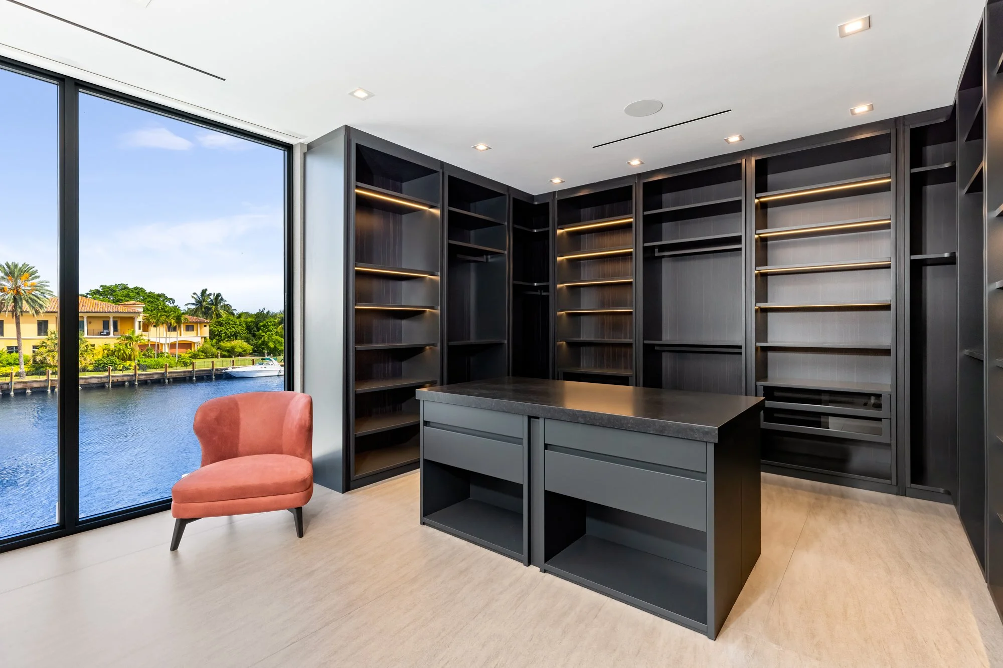 Modern home office with black built-in bookshelves, a black island table, a pink upholstered chair, and large windows overlooking a body of water and houses with palm trees.