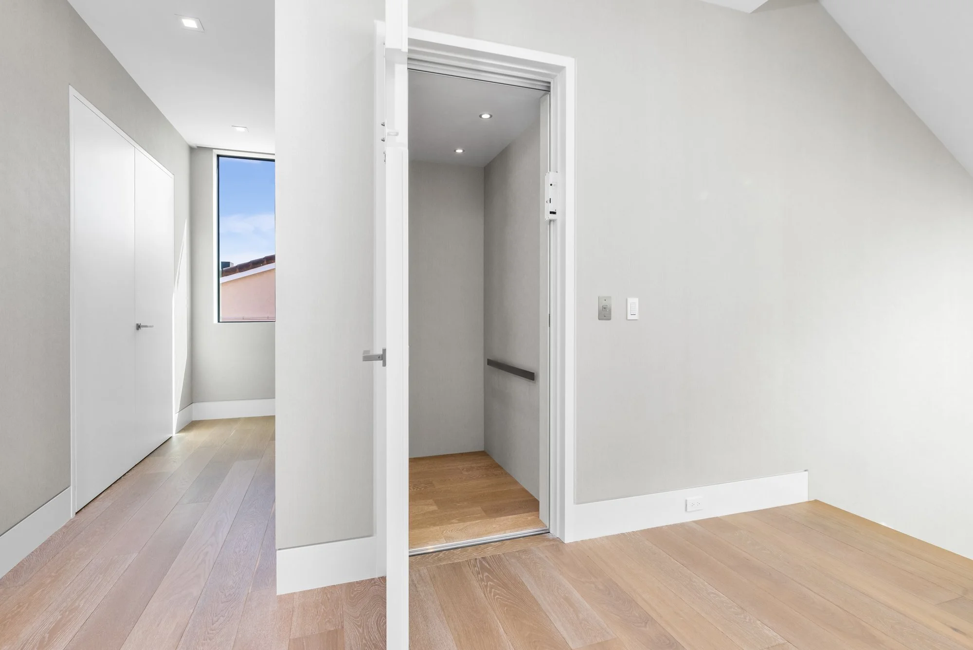 Empty room with light-colored walls, wooden floor, a door to an elevator, a window showing part of a roof and sky, and a white wall with a power outlet.