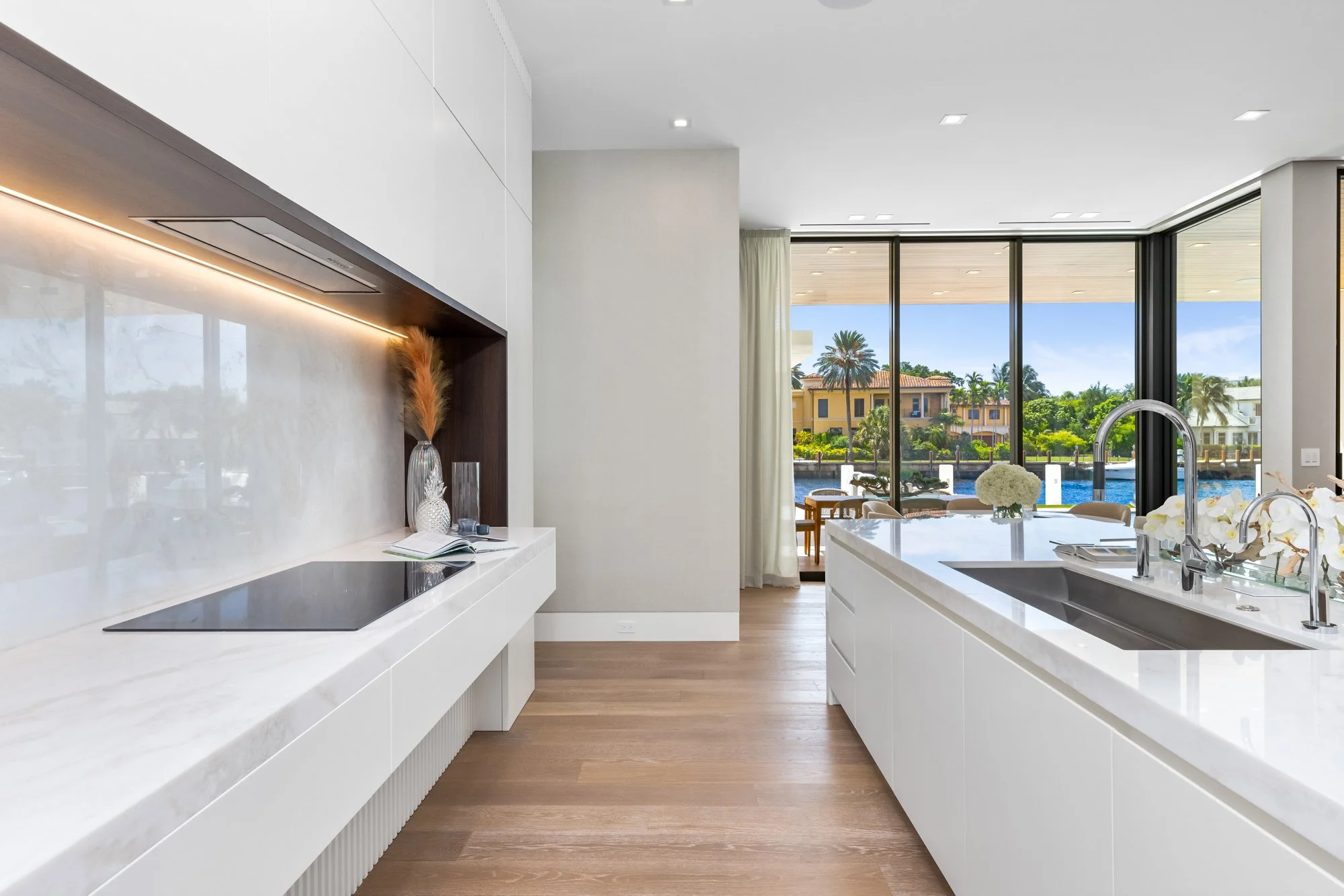 Modern kitchen with white cabinetry, marble countertops, and large windows overlooking a canal with houses and palm trees.