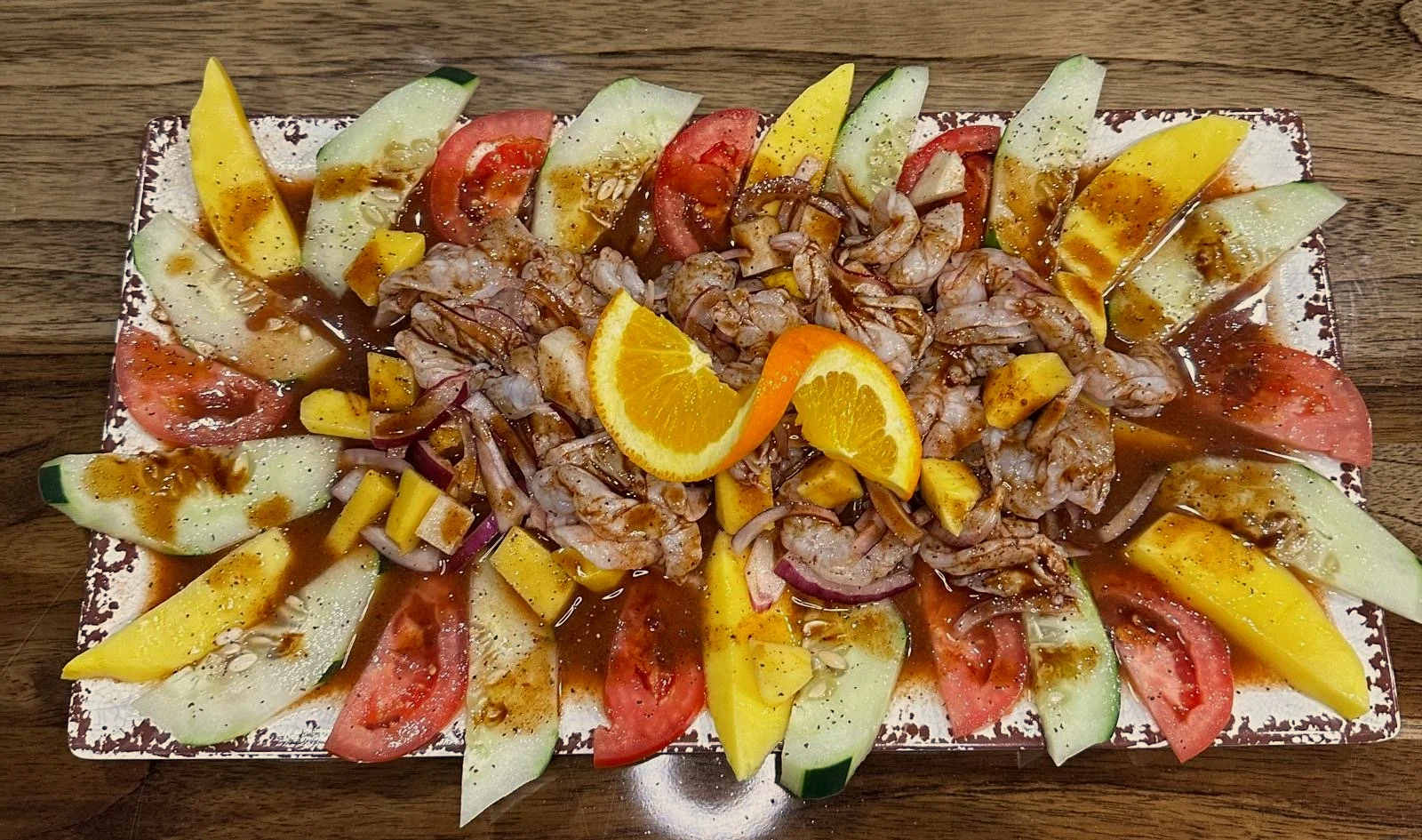 Seafood platter with sliced tomatoes, cucumbers, mango, and oranges, topped with seafood, sauce, and garnished with lemon slices.