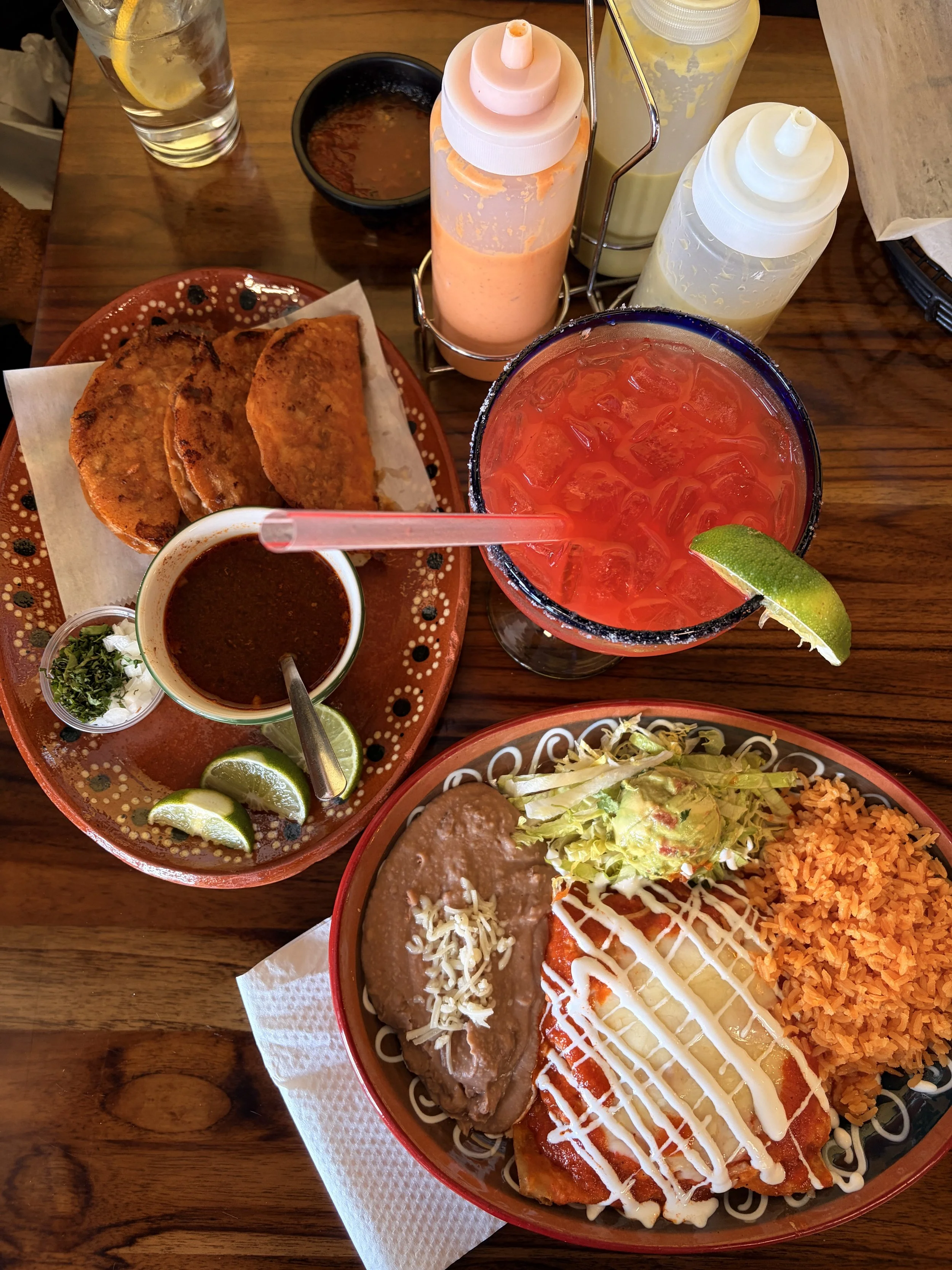 A Mexican meal with fried tacos, rice, refried beans with cheese, shredded lettuce, and a drizzled white sauce, along with a large red fruit drink with lime wedge, two plates of liquid condiments, and two squeeze bottles of sauce on a wooden table.