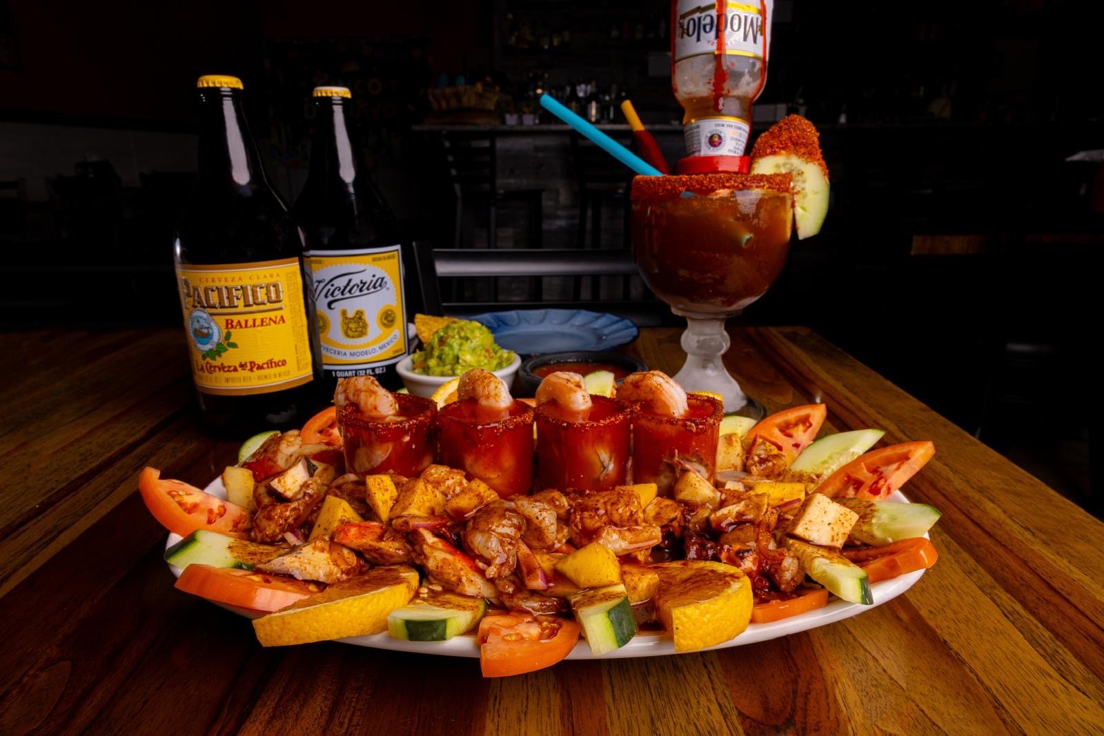 A plate of Mexican food with stuffed peppers, surrounded by sliced vegetables, with a side of guacamole, and drinks including two bottles of beer and a glass of Michelada with garnishes on a wooden table.