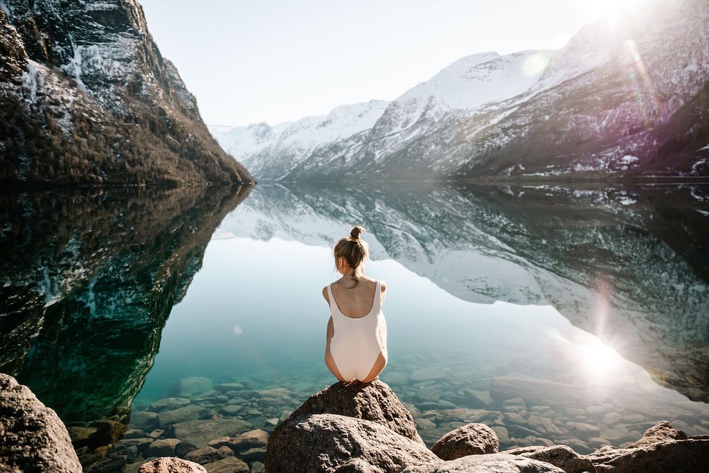 En kvinne sitter på en stor stein ved en rolig fjord omgitt av snødekte fjell, med sollys som reflekteres i vannet.