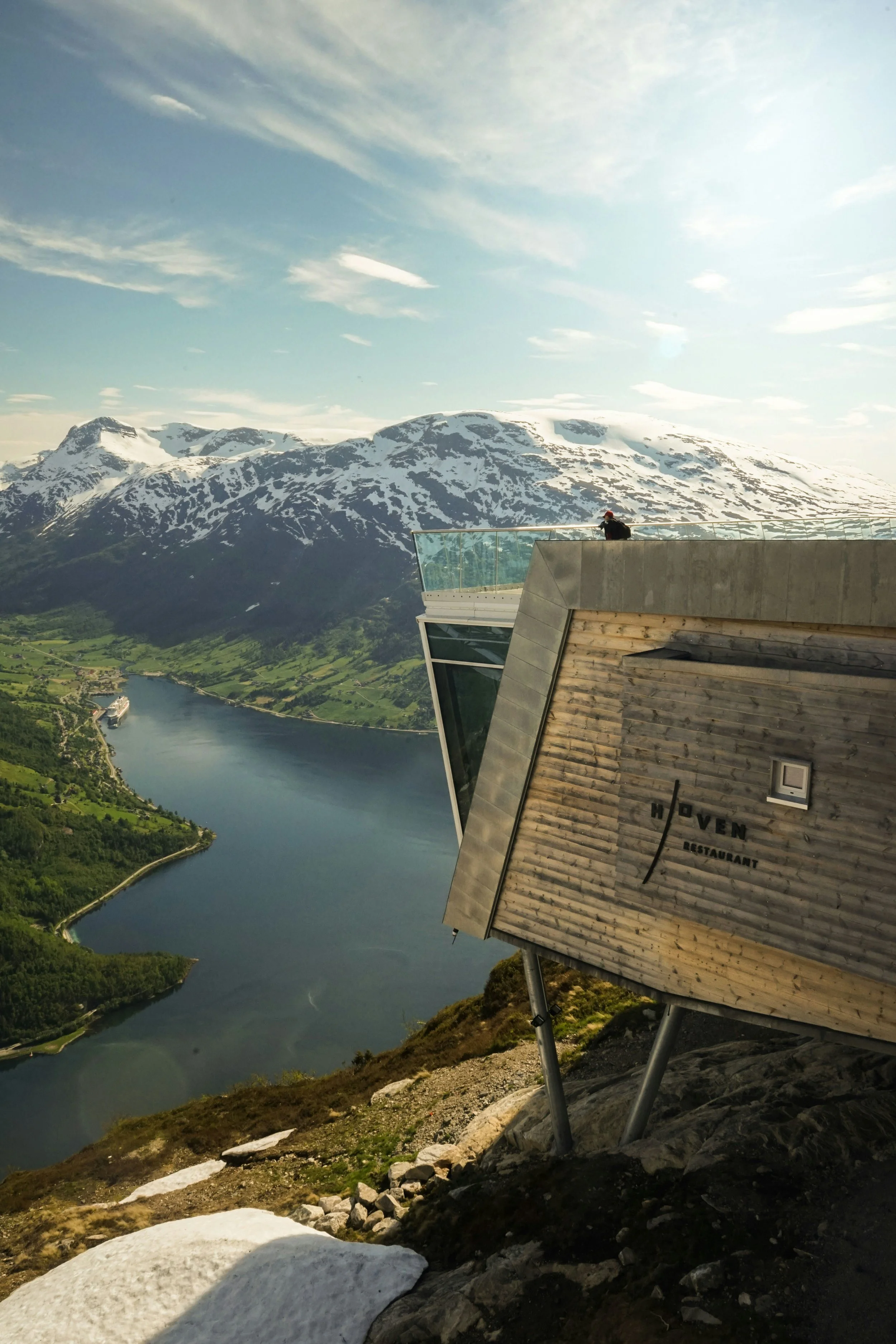 Et moderne restaurantbygg med en person som står på en uforglemmelig utsiktsplattform, med snødekte fjell og en innsjø i bakgrunnen, omgitt av grønt landskap.