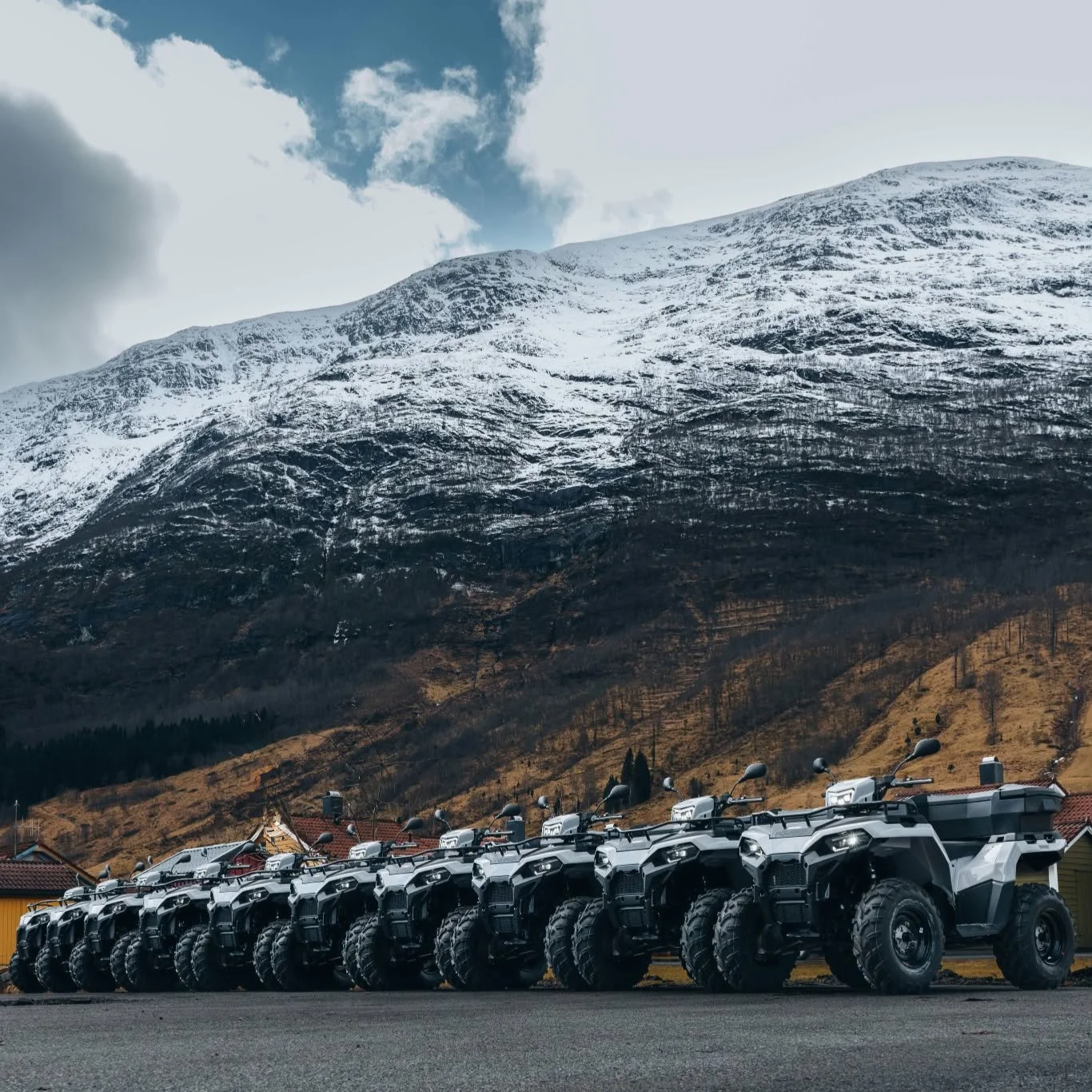 En rekke med hvite og svarte ATV-er parkert foran en bygning med fjell i bakgrunnen, snø på toppen av fjellene og en blå himmel med noen skyer.