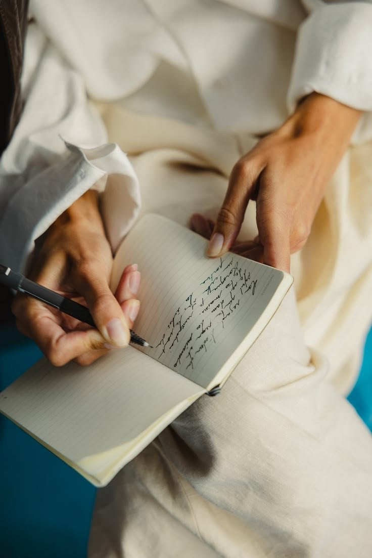 Person writing in a notebook with a black pen, wearing a white long-sleeve shirt and beige pants, seated on a blue surface.