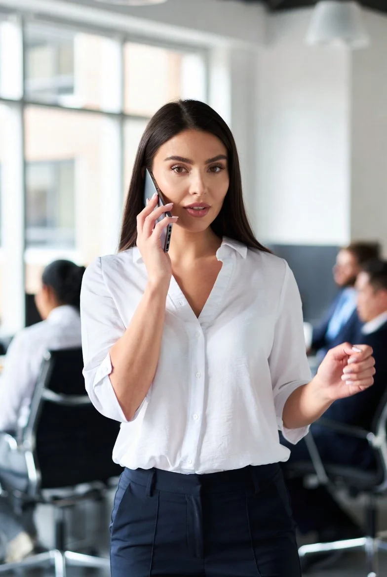 A woman with long dark hair in a white shirt talking on a mobile phone in an office setting with coworkers in the background.