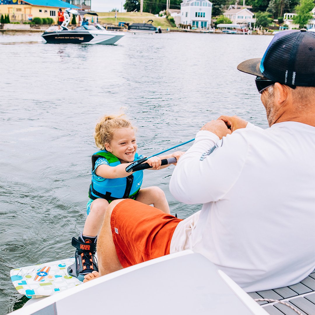 A young girl with curly blonde hair sitting on a paddleboard, wearing a life jacket and holding a fishing pole, smiling at an adult man sitting in a boat on a lake, with houses and boats visible in the background.