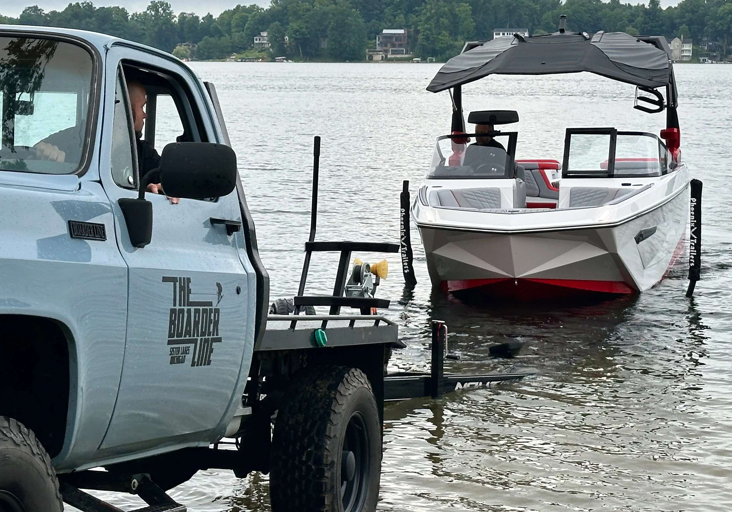 A boat is being launched into the water from a trailer attached to a small truck on a lakeshore. The boat is white with a black canopy, and it has red accents inside. The truck has a logo that reads 'The Border Life' and is parked near the water's edge.