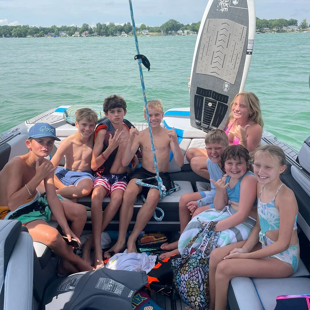 Group of children and a woman on a boat, enjoying a day on the water, with a wakeboard leaning against the side of the boat.
