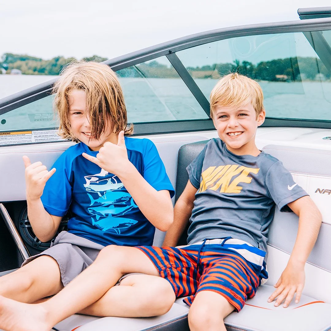 Two young boys sitting on a boat's seat, boat in the background, one making a shaka hand sign and the other smiling.