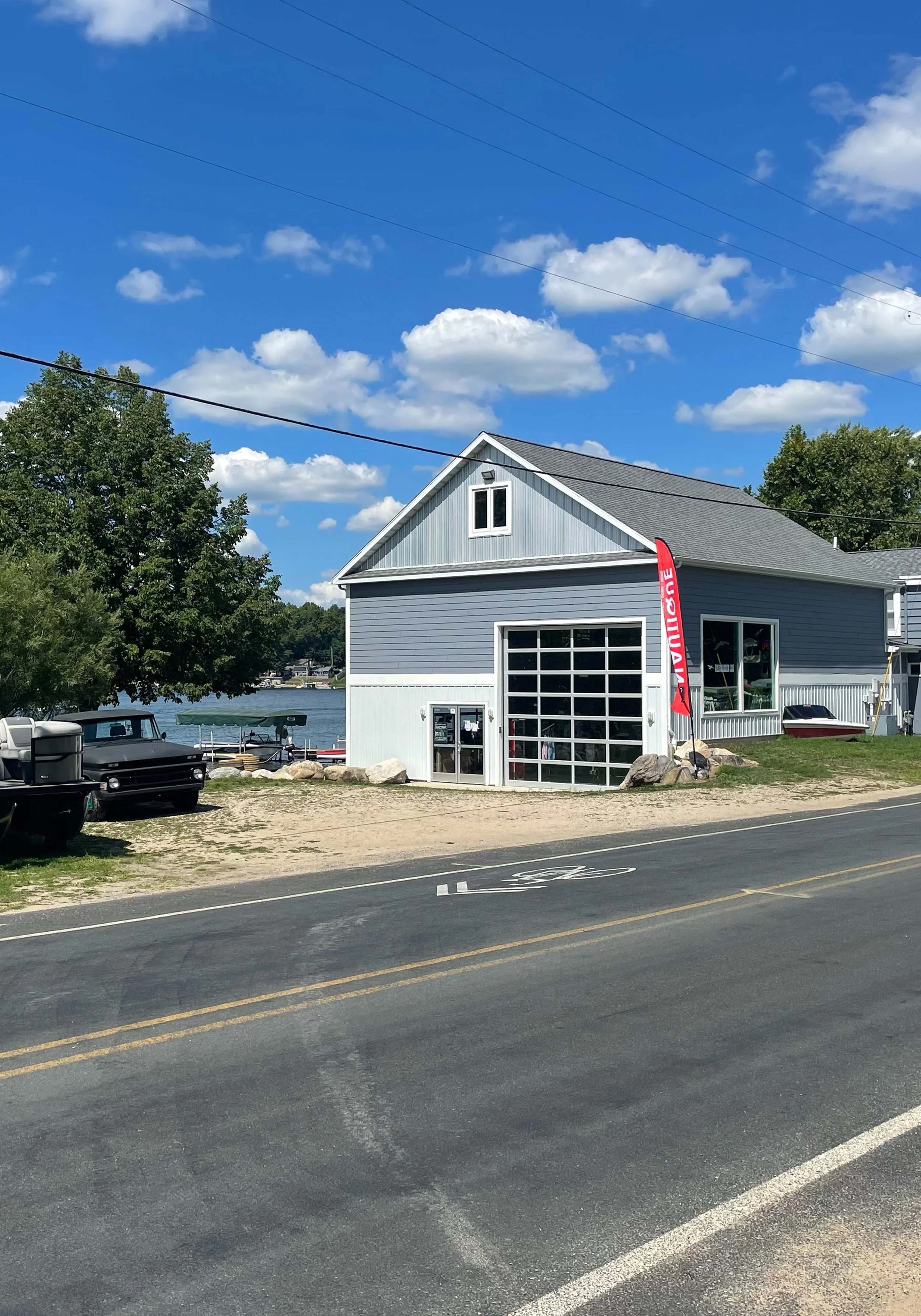 A waterfront building with a large garage door, a red flag with white text, parked cars, a body of water, trees, and a bright blue sky with clouds.