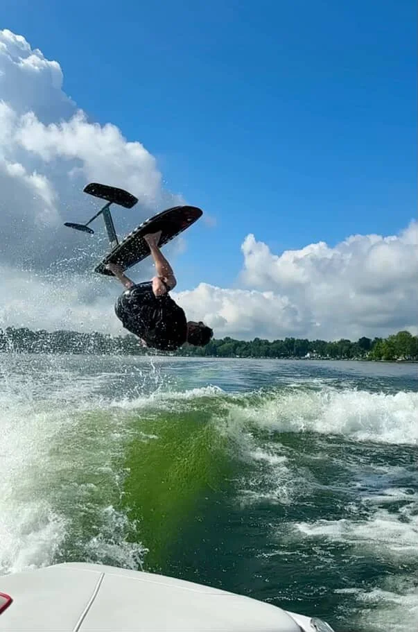 Person flipping upside down on a wakeboard in the water on a sunny day with blue sky and clouds.