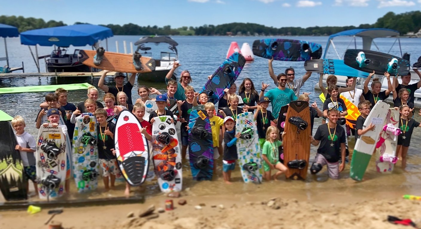 A large group of children and adults holding wakeboards and stand-up paddleboards on a sandy beach near a body of water, smiling and celebrating.