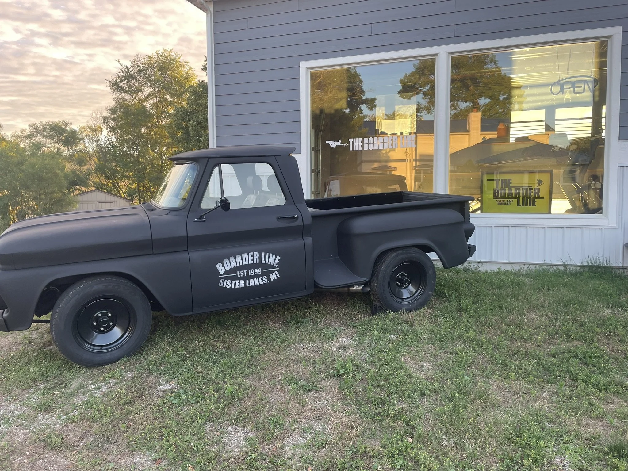 A black pickup truck parked on grass in front of a building with large glass windows. The windows display signs for 'The Boarder Line' in Sister Lakes, MI, with an open sign visible inside. Trees and a partly cloudy sky are reflected in the windows, and the scene appears to be at sunset.