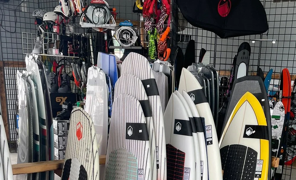 Surfboards displayed upright in a retail store, along with helmets, bags, and other sports gear on a wire grid display.