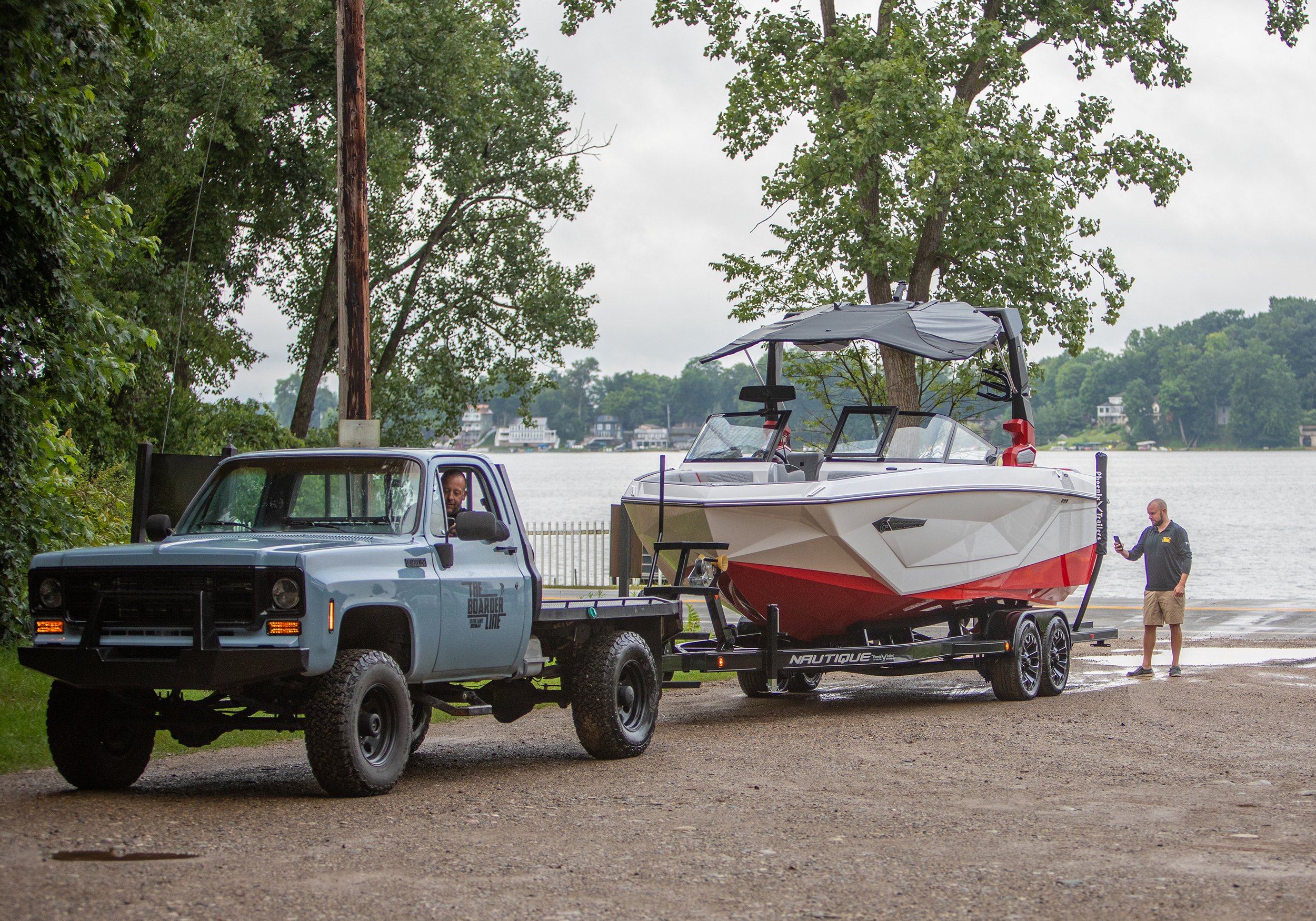 A pickup truck parked near a lake with a boat on a trailer, and two men near the boat, one inside the truck and the other standing on the ground