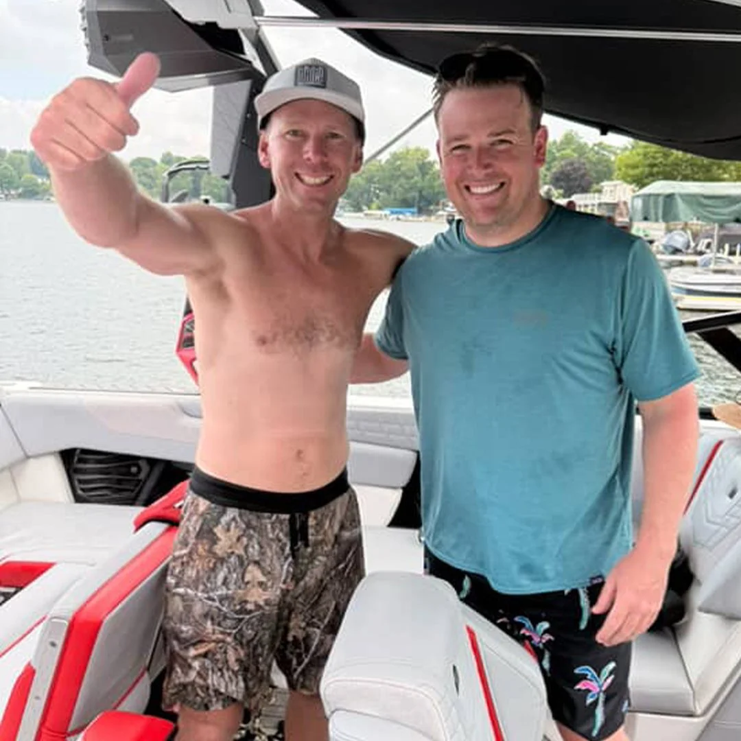 Two men enjoying time on a boat; one shirtless, wearing camouflage shorts and a baseball cap, giving a thumbs-up; the other in a blue t-shirt and patterned shorts, smiling; water and boats visible in the background.