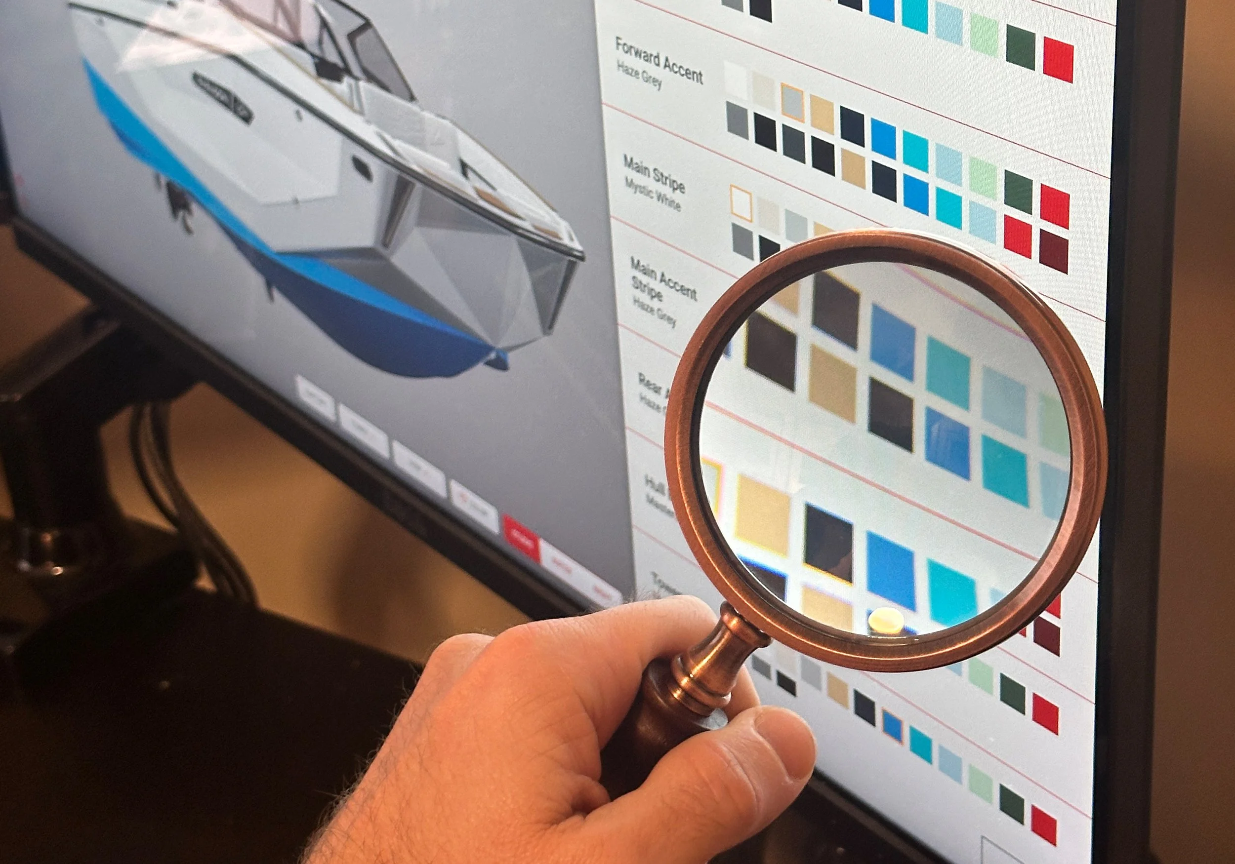 Person inspecting a design on a computer screen with a magnifying glass, which shows color swatches and a boat graphic.
