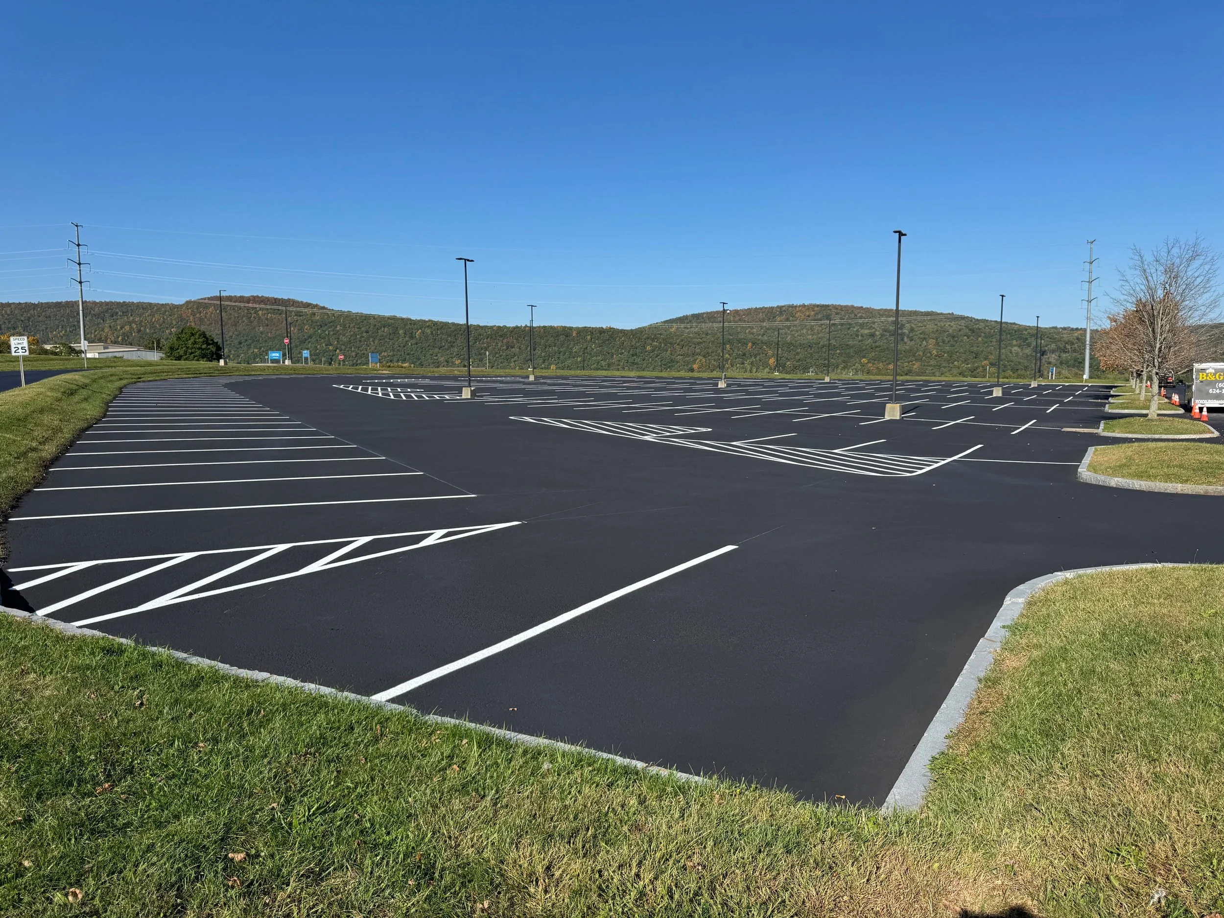 Empty parking lot with freshly paved black asphalt, white painted parking lines, lamp posts, green grass, and a backdrop of distant hills under a clear blue sky.