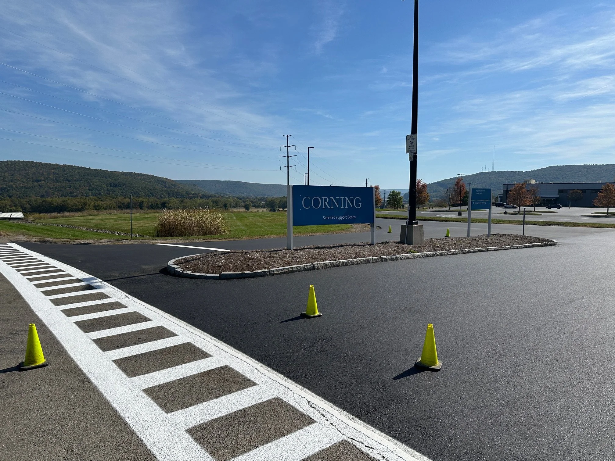 Parking lot with yellow traffic cones, a blue sign that says 'Corning Services Support Center', and a large empty grassy area with hills in the background.