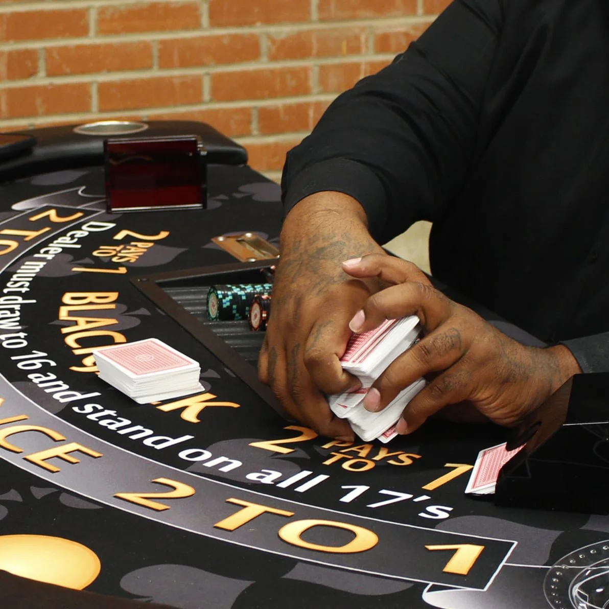 A person shuffles playing cards at a blackjack table, with poker chips and a brick wall in the background.