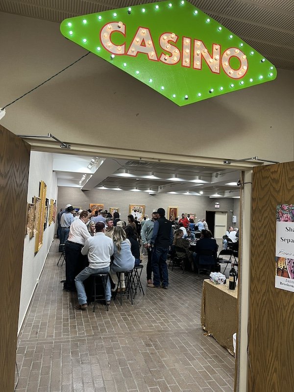 People gathered inside a casino event, with some sitting at tables and others standing, in a well-lit room decorated with artwork on the walls.