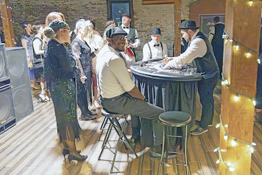 Group of people at a music performance, some standing and listening, one person playing a piano, in a decorated indoor venue.