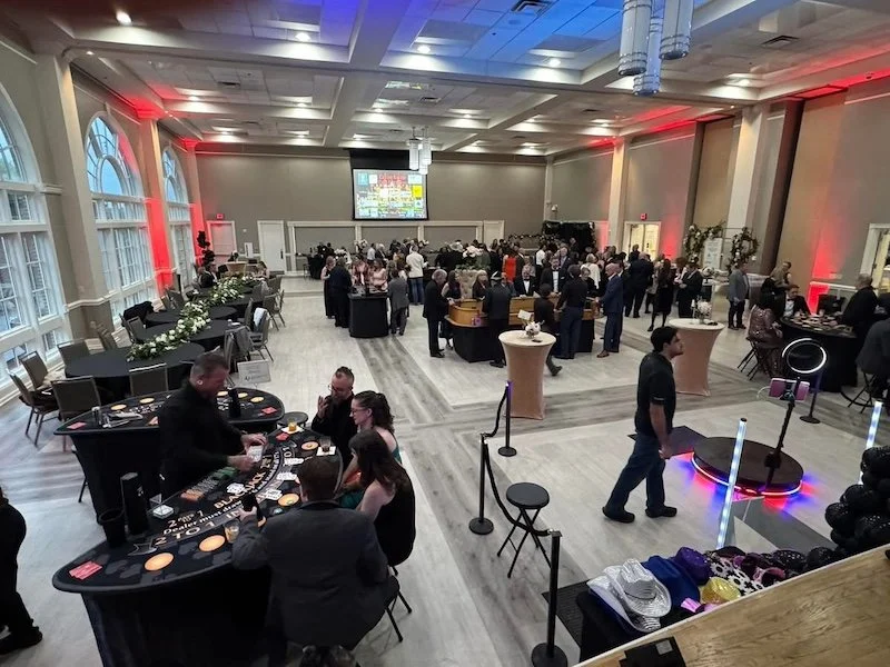 Large indoor event with people mingling, buffet tables, a photo booth, and a projection screen, in a decorated banquet hall with large windows and red accent lighting.