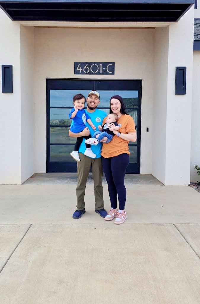 Family of four standing in front of a house with the number 4601-C above the door. The family includes a man, woman, and two children, all smiling and holding the kids.